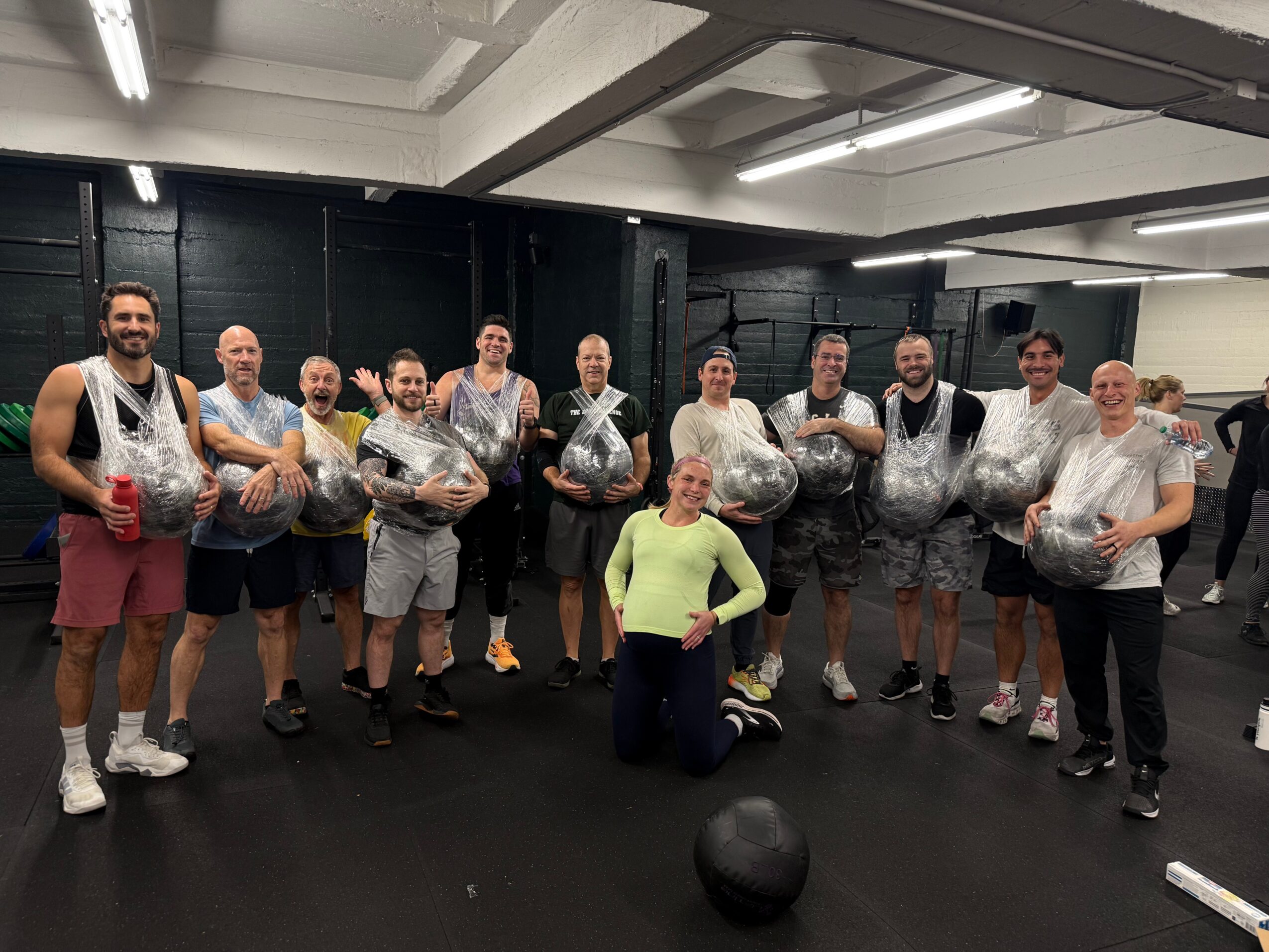 Group of people in a gym holding large bags, some wearing workout attire, with a woman kneeling in front, a black ball on the floor.