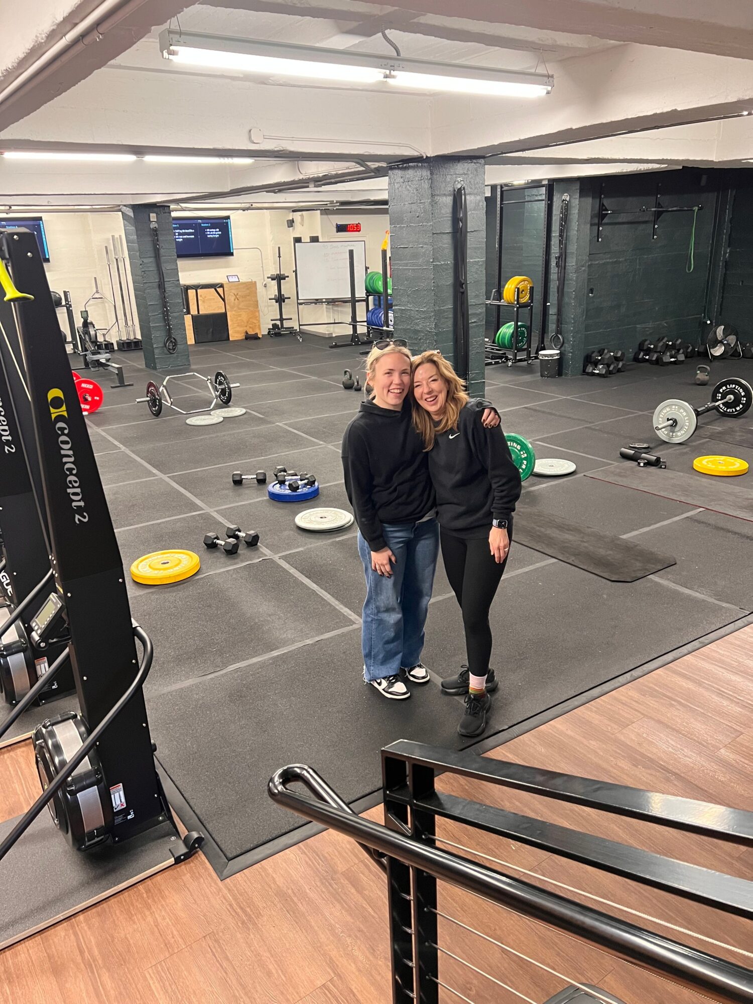 Two women in a gym, one with blonde hair and the other with dark hair, standing near gym equipment and weights.