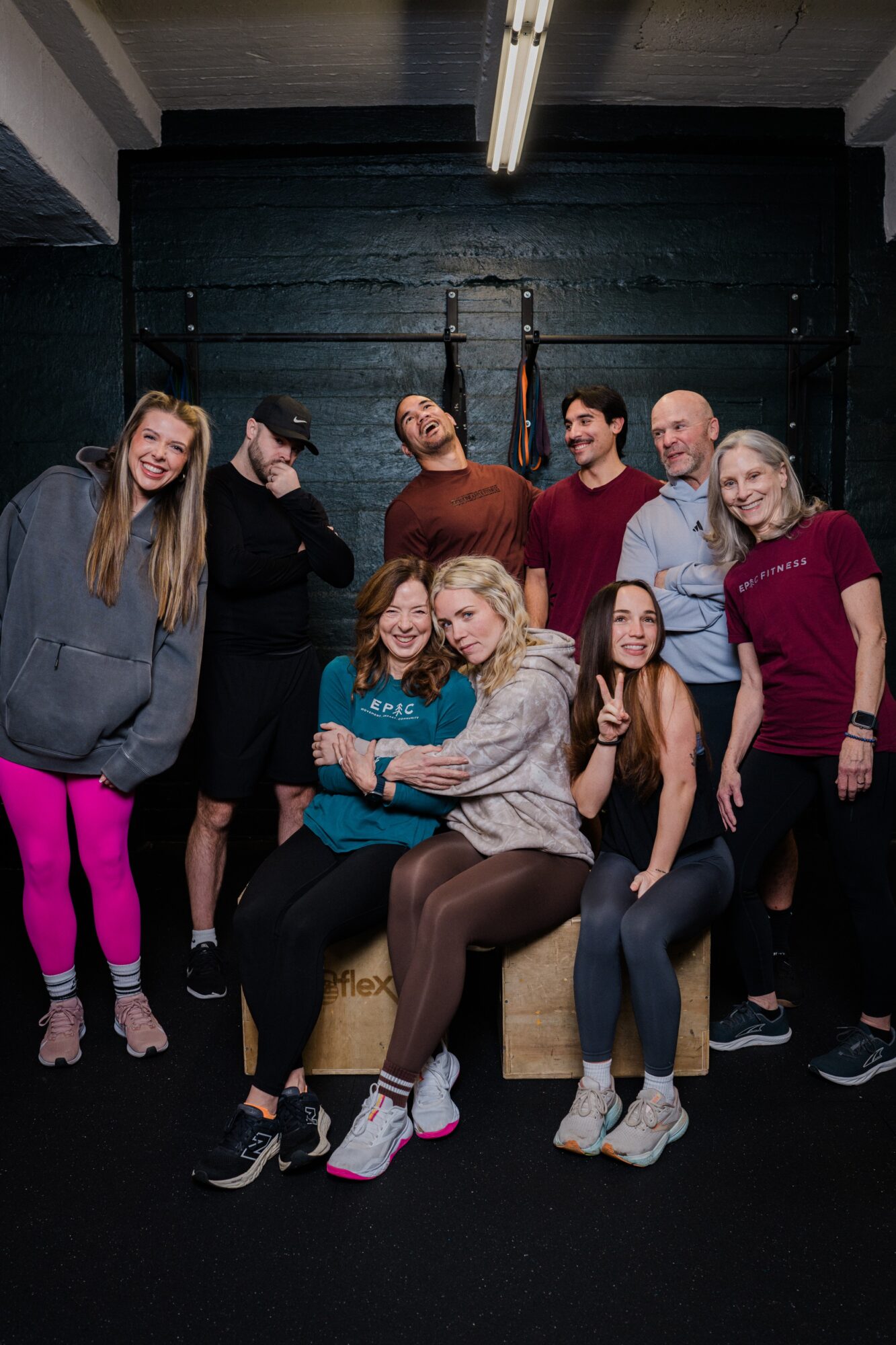 Group of nine people sitting and standing in a dark room with a black wall and overhead lighting, smiling and posing.