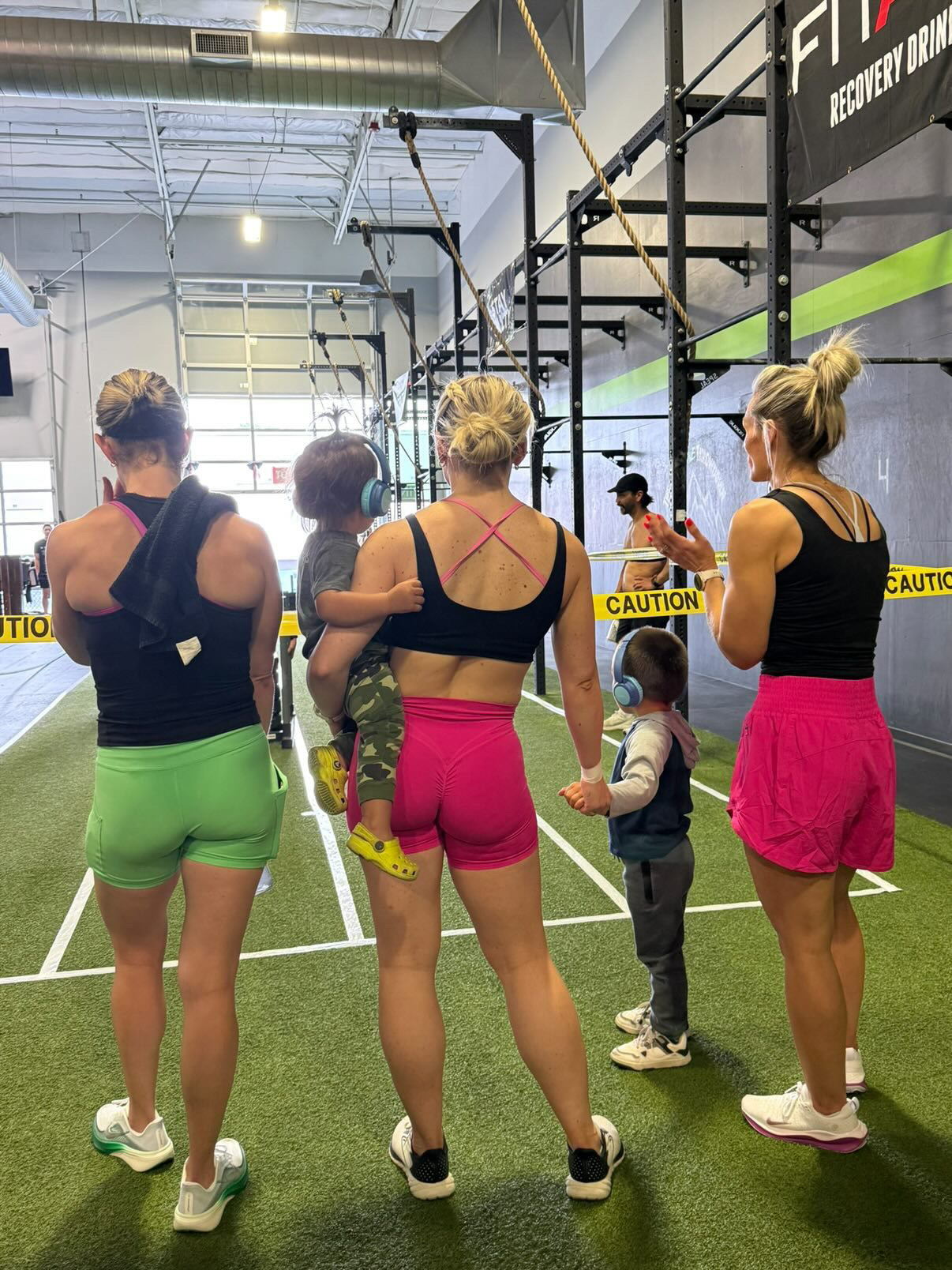 Four women and a girl stand in a gym with black equipment and green turf, facing away, with sunlight coming through windows.