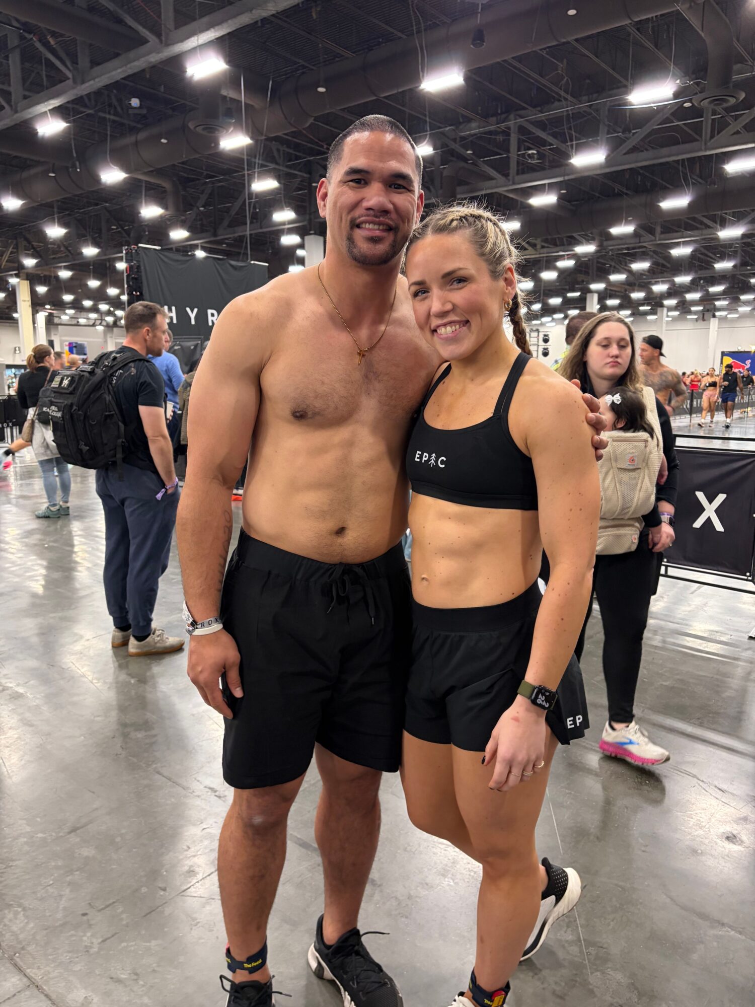 A shirtless man and a woman in athletic wear pose together at an indoor event, with people and booths in the background.