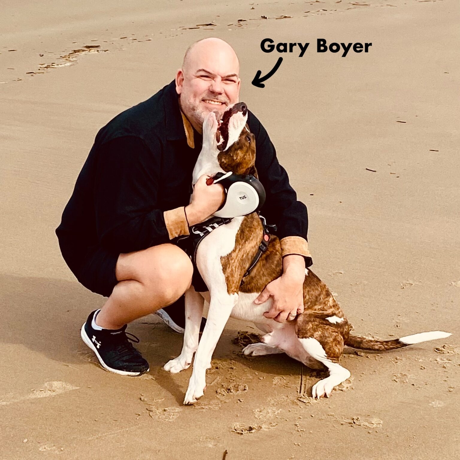 Person crouching on sand, smiling, with a dog licking their face, labeled Gary Boyer, on a beach.