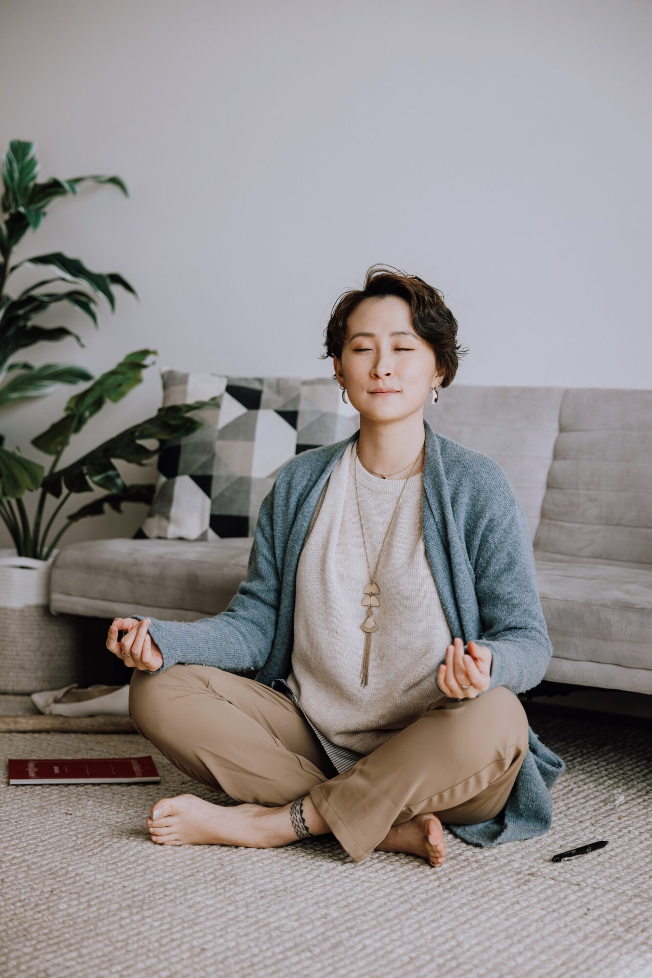 Person sitting cross-legged on the floor in a meditative pose, eyes closed, in a living room with a sofa and plant