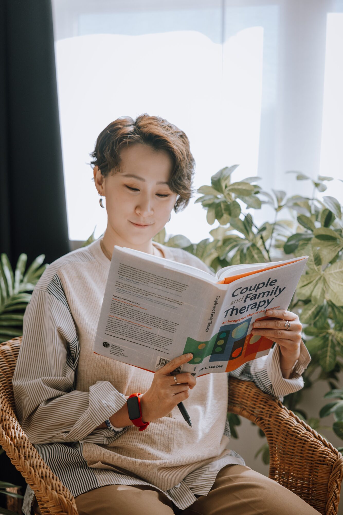 Person with short hair reading a book in a room with plants and natural light, sitting on a wicker chair.