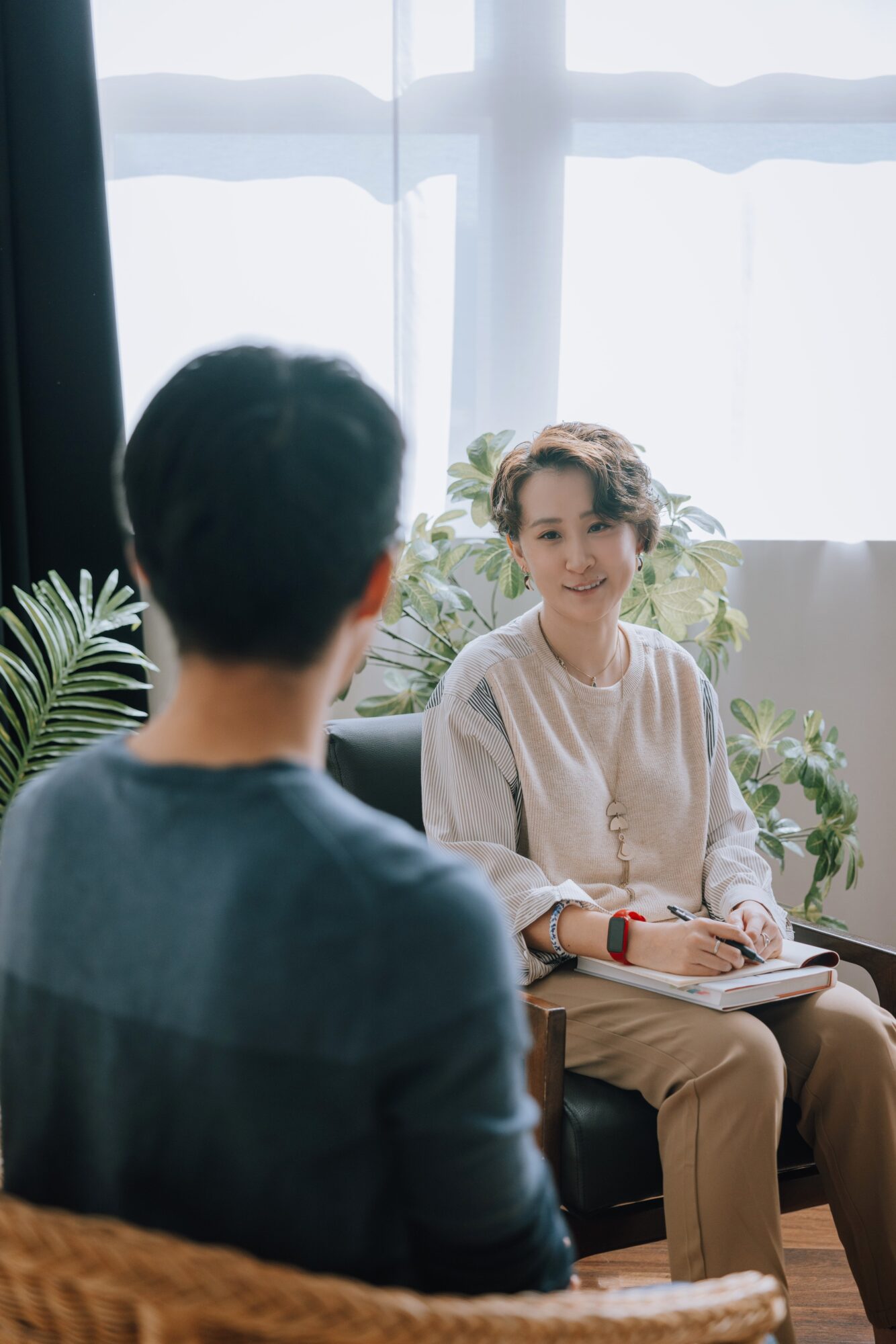 Woman smiling and talking to a man in a room with large windows and plants, holding a notebook.