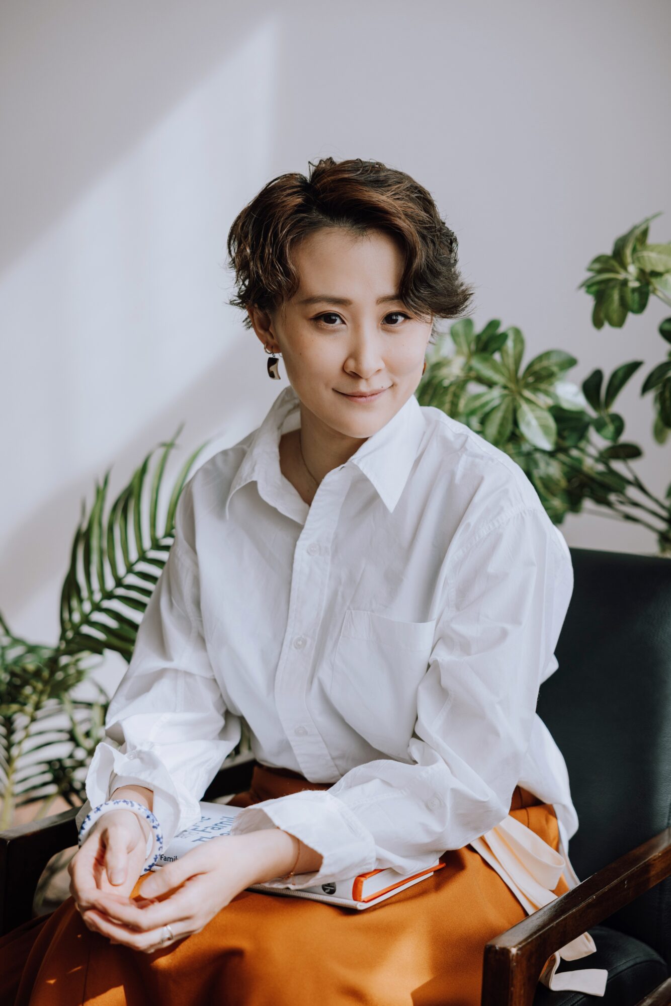 Woman with short curly hair wearing a white shirt, sitting in front of green plants, smiling at the camera.