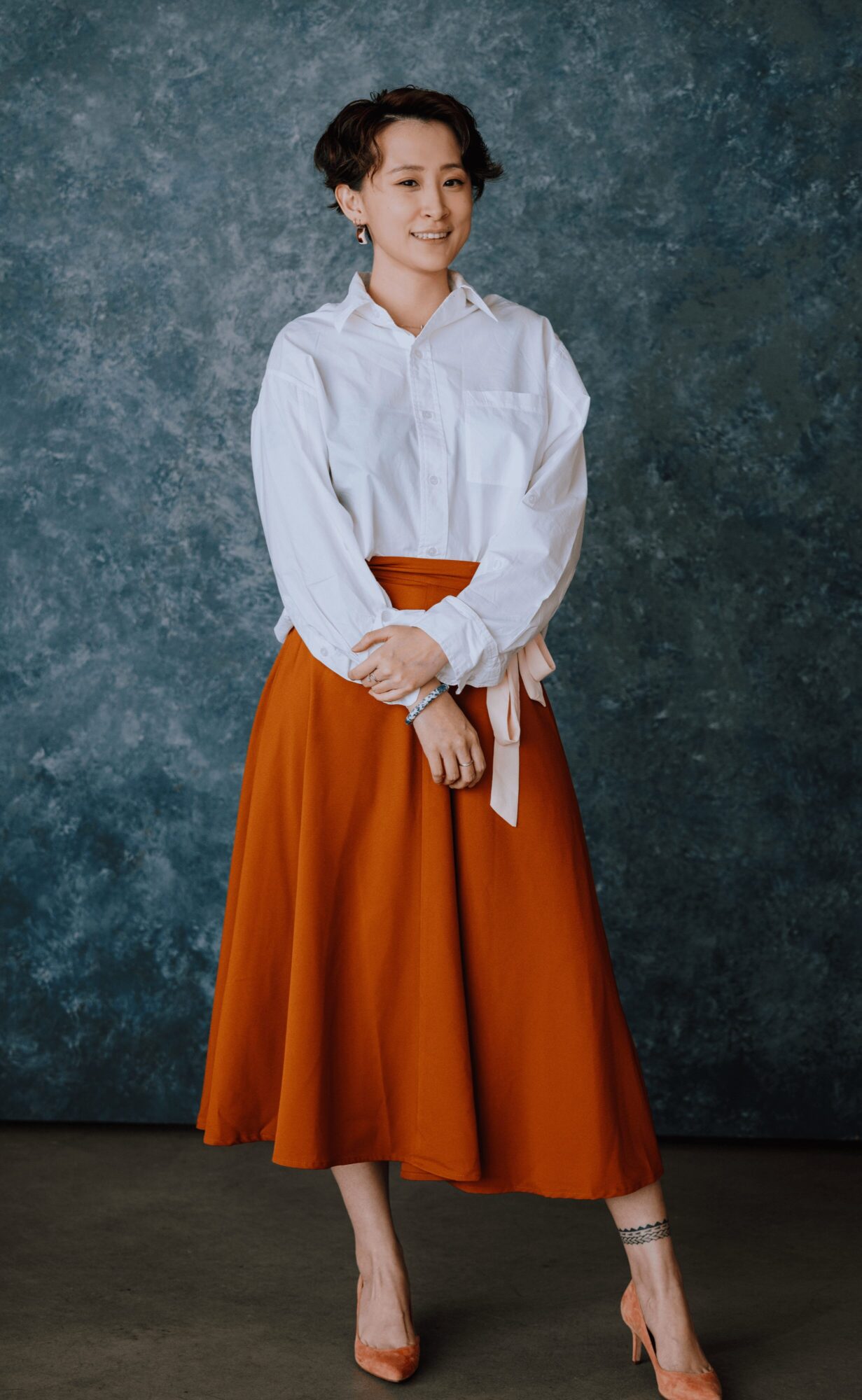 Young woman in white blouse and orange skirt standing against textured blue background, smiling, with hands clasped.