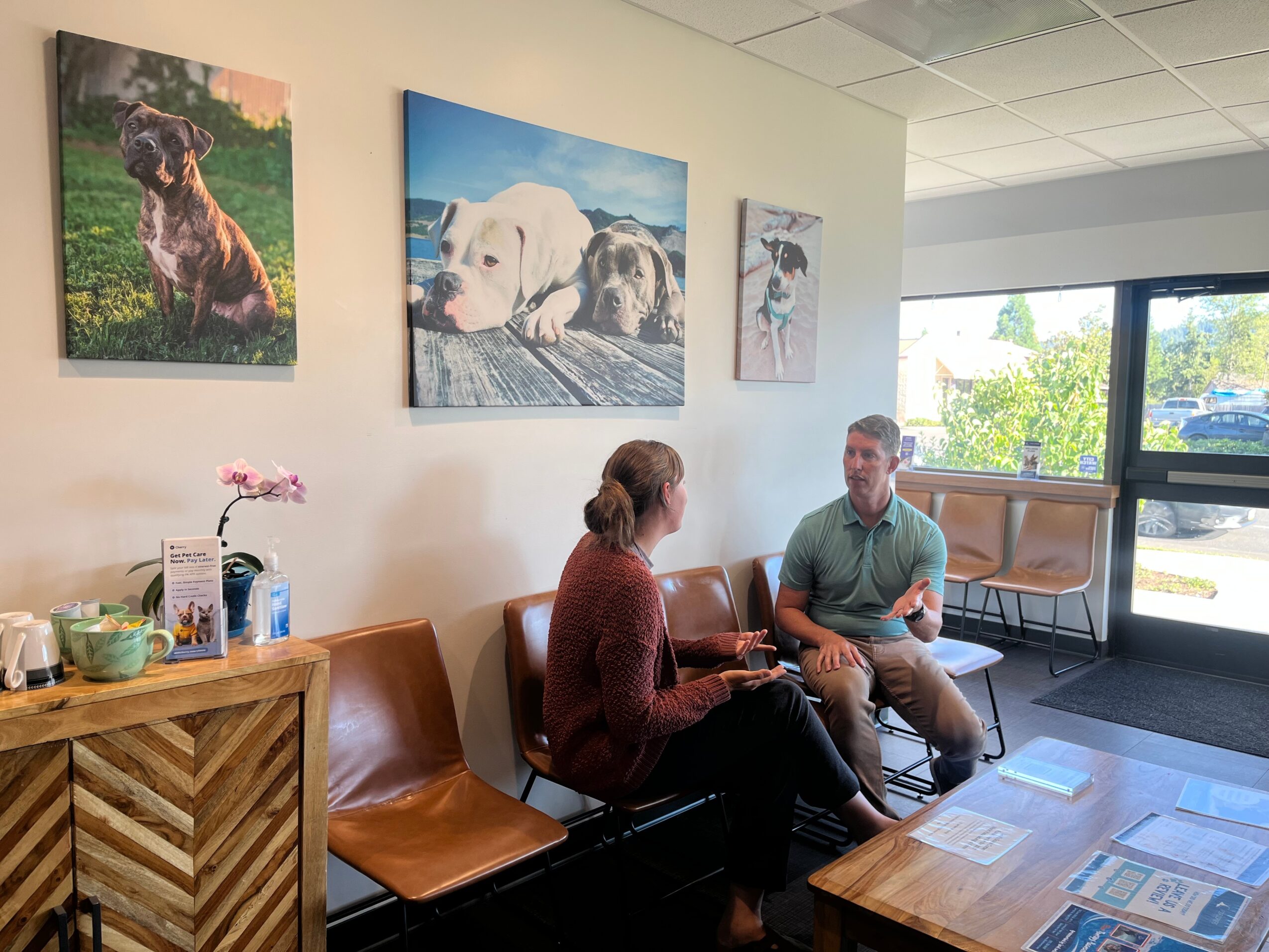 Two people sit on a bench in a room with dog paintings on the wall, a window showing outside greenery, and a wooden table.