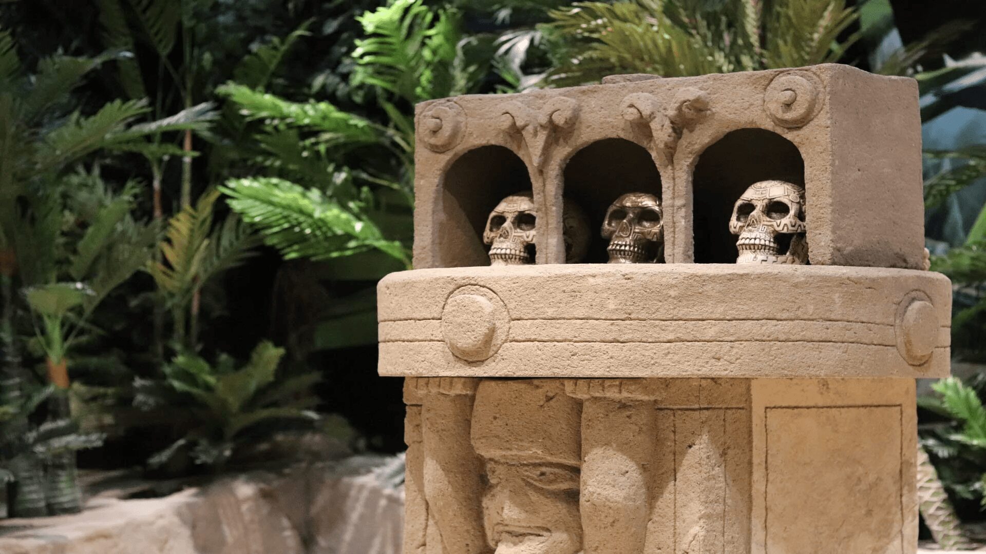 Stone structure with three skull carvings in arched niches, surrounded by lush green plants.