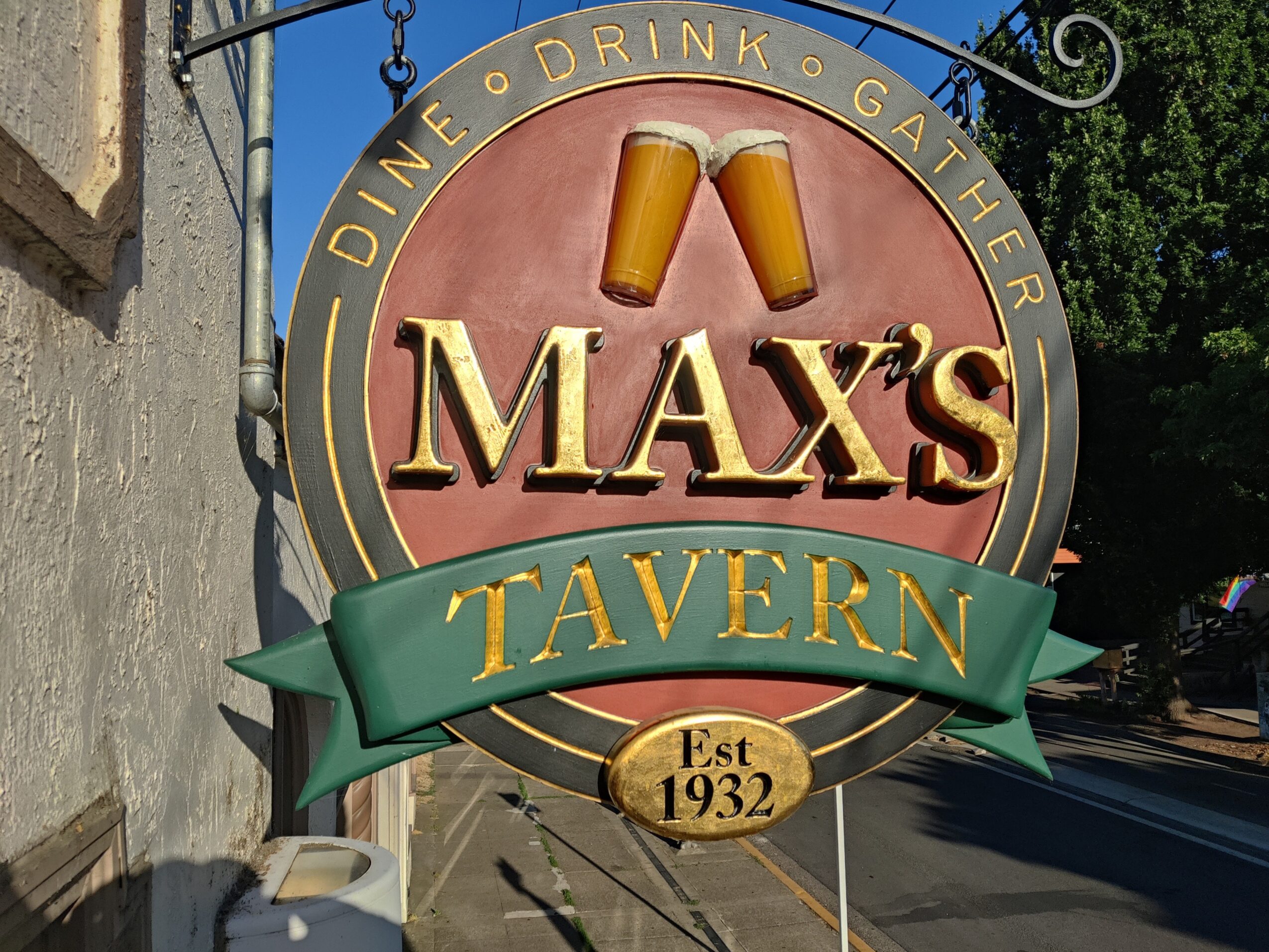 Sign for Max's Tavern with two beer glasses, established 1932, on a street corner, with trees and a building nearby.