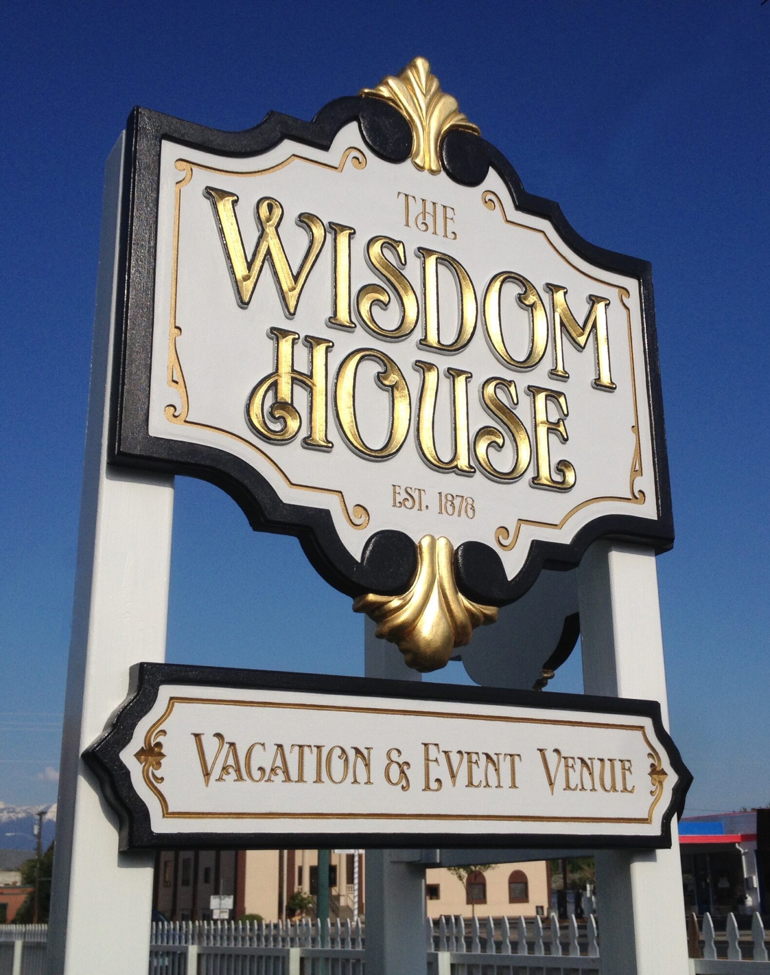 Sign for The Wisdom House, vacation and event venue, with gold decorative elements and clear text, against a blue sky.