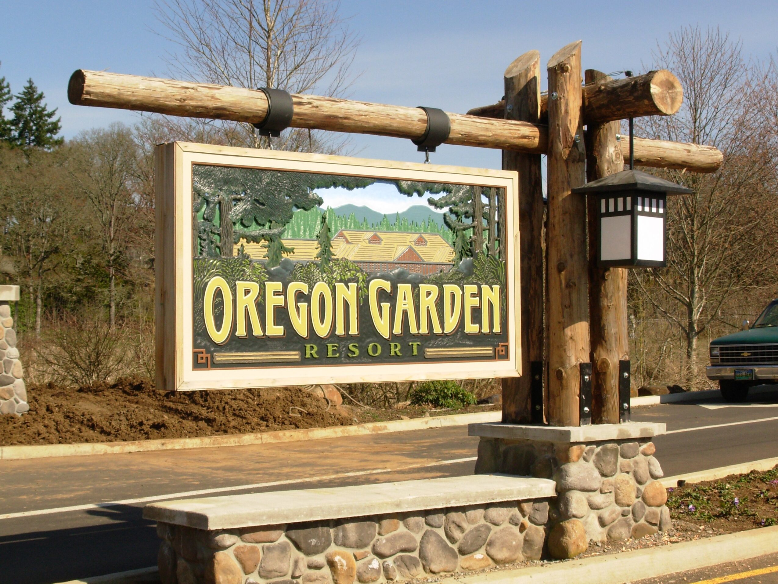 Sign for Oregon Garden Resort with wooden posts and a stone base, set against a background of trees and a road.
