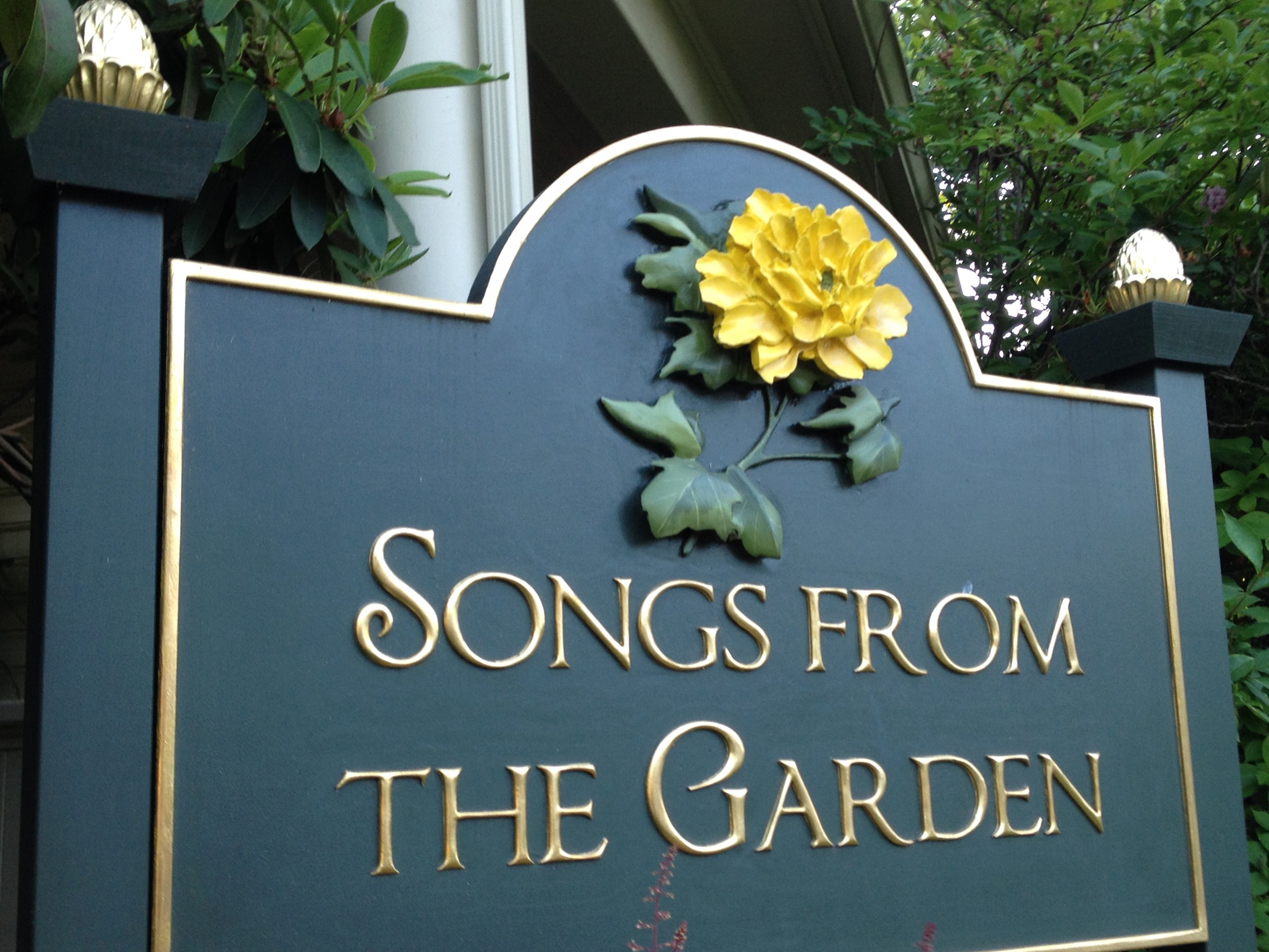 Sign with a yellow flower and green leaves, reading 'Songs from the Garden' in gold letters.