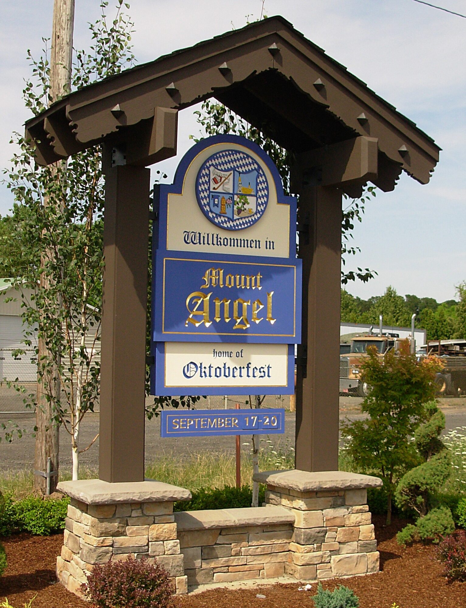 Welcome sign for Mount Angel, featuring a blue and white design with a church and a cross, on a wooden structure with stone base.