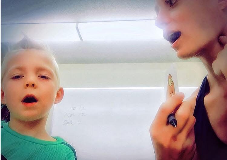 Child and adult in a classroom, child with mouth open, adult holding a pen near face, whiteboard in background.