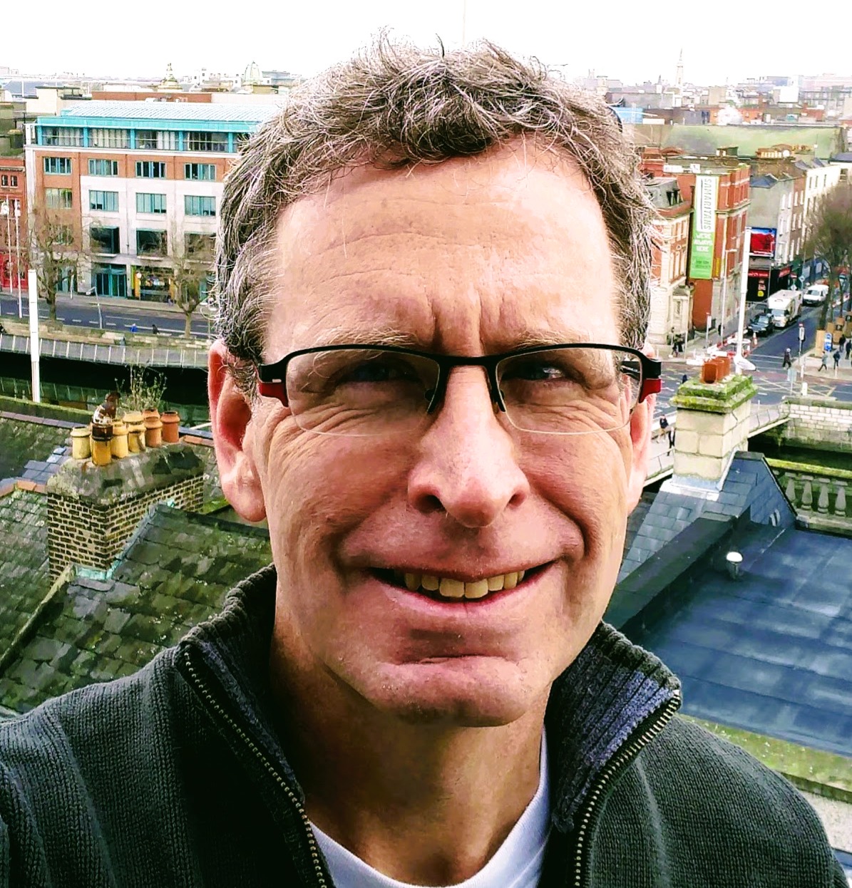 Man with glasses smiling outdoors, cityscape background, buildings, and streets visible behind him.