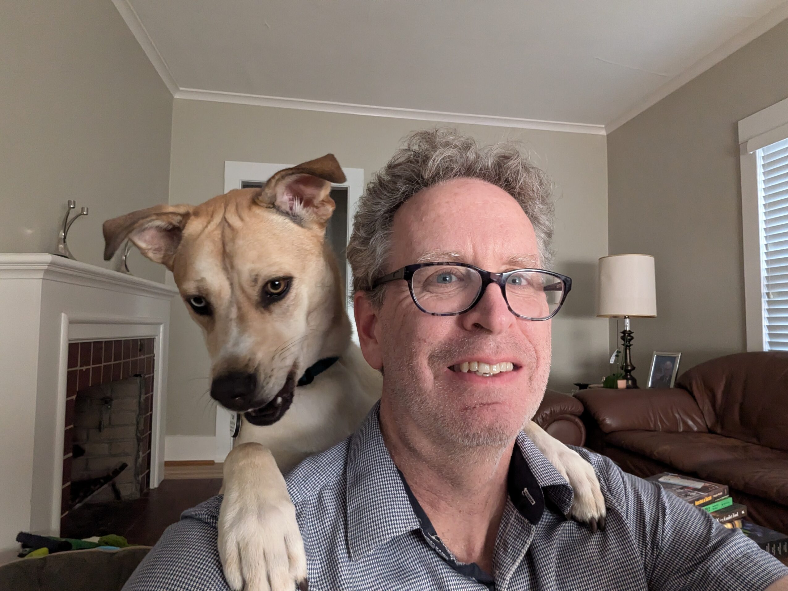 A man with glasses smiling, with a dog resting its paw on his shoulder in a living room.