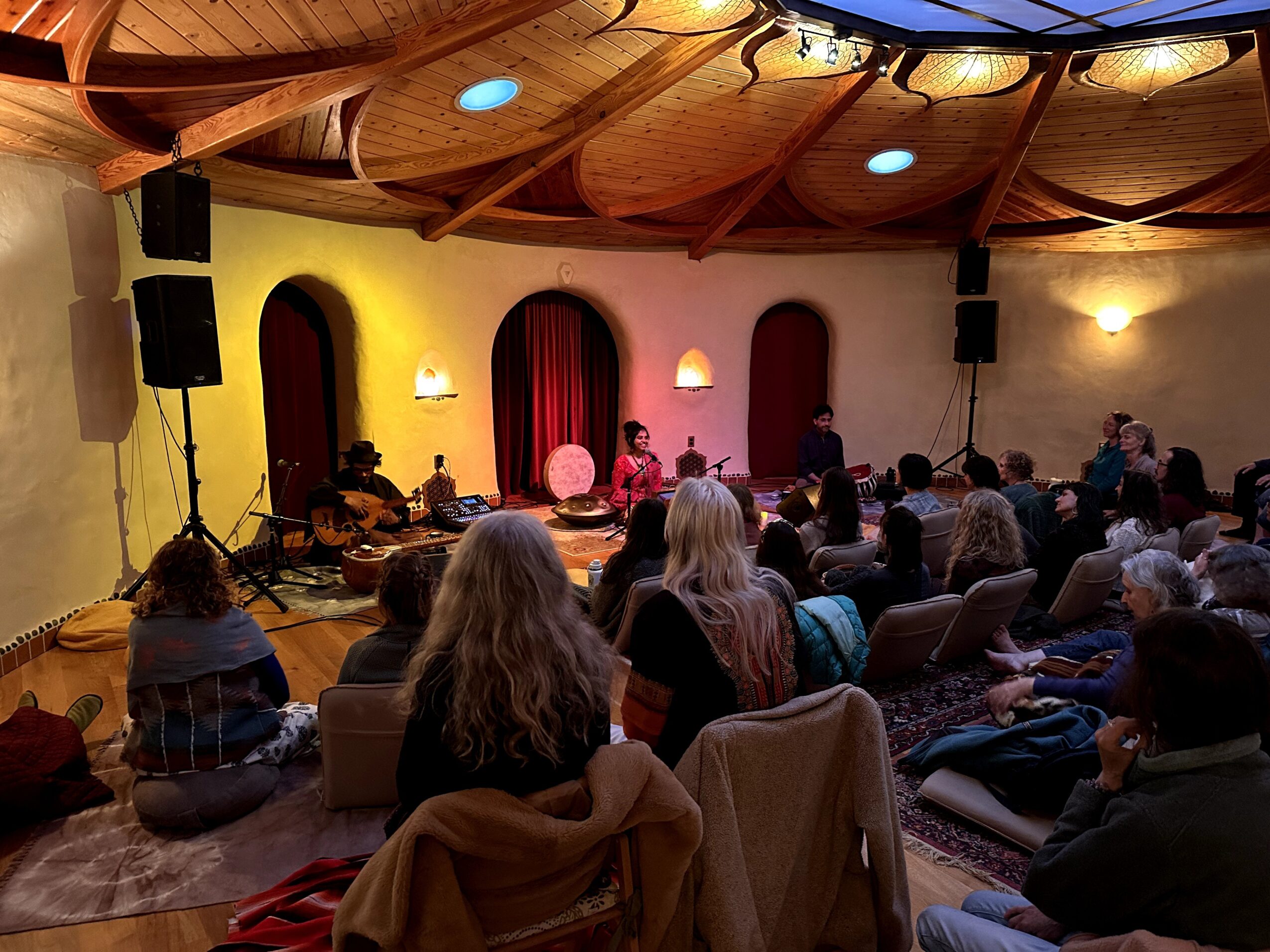 Audience seated on cushions and chairs in a room with a wooden ceiling, watching a performance on stage with red curtains.