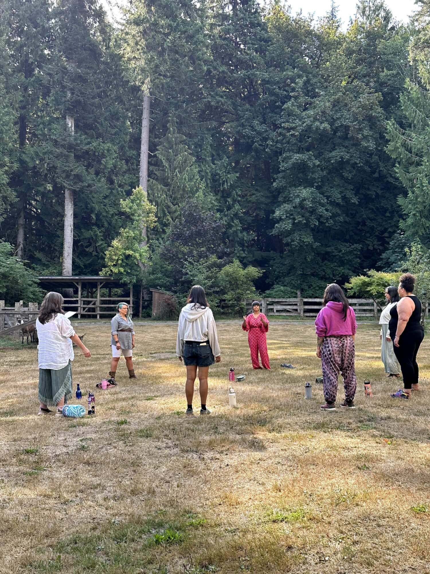 Group of people standing outdoors on grass with trees in the background, spaced apart, some facing forward, some turned sideways.