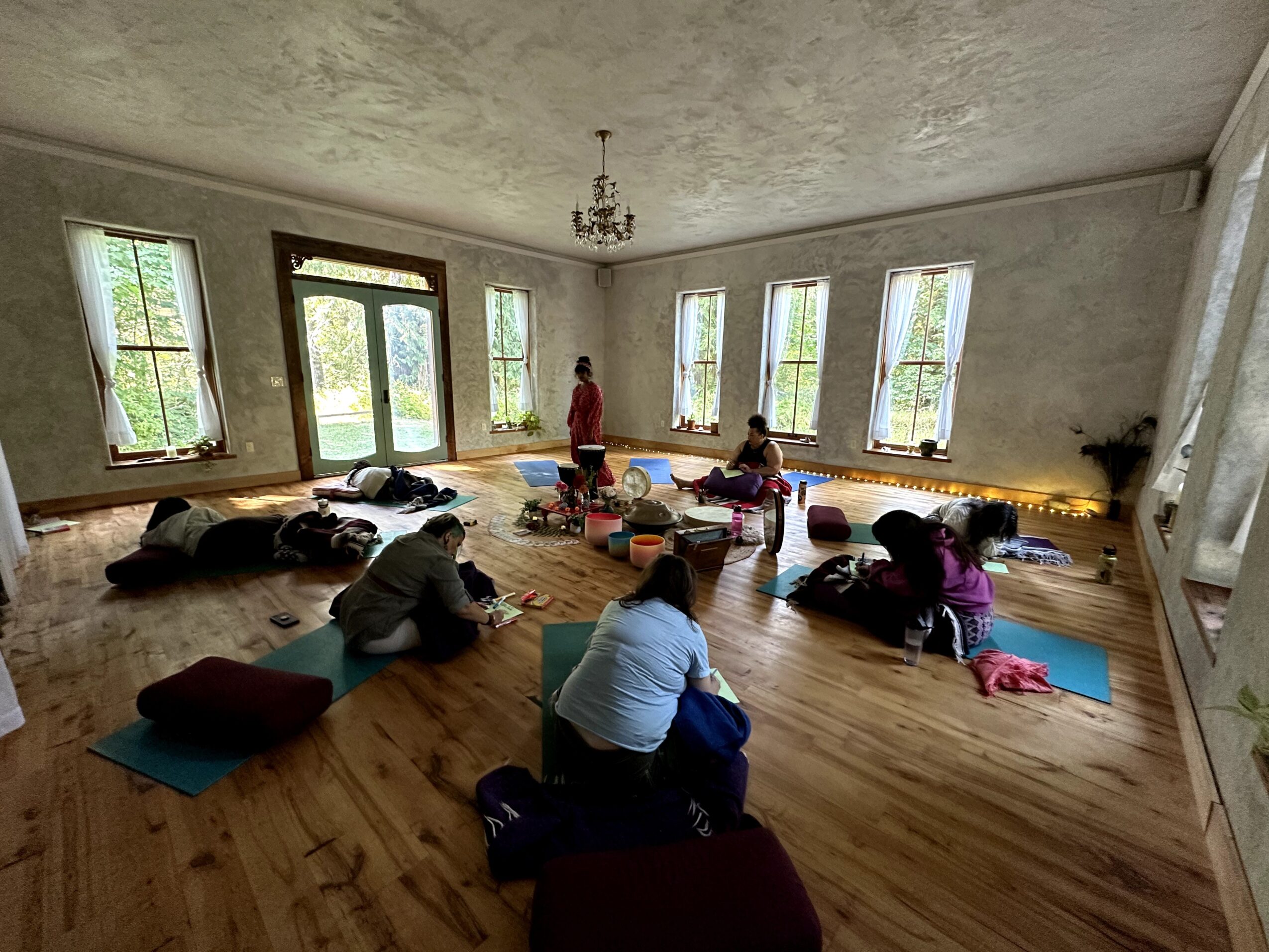People sitting on cushions in a spacious room with large windows, practicing yoga or meditation, with a person standing at the front.