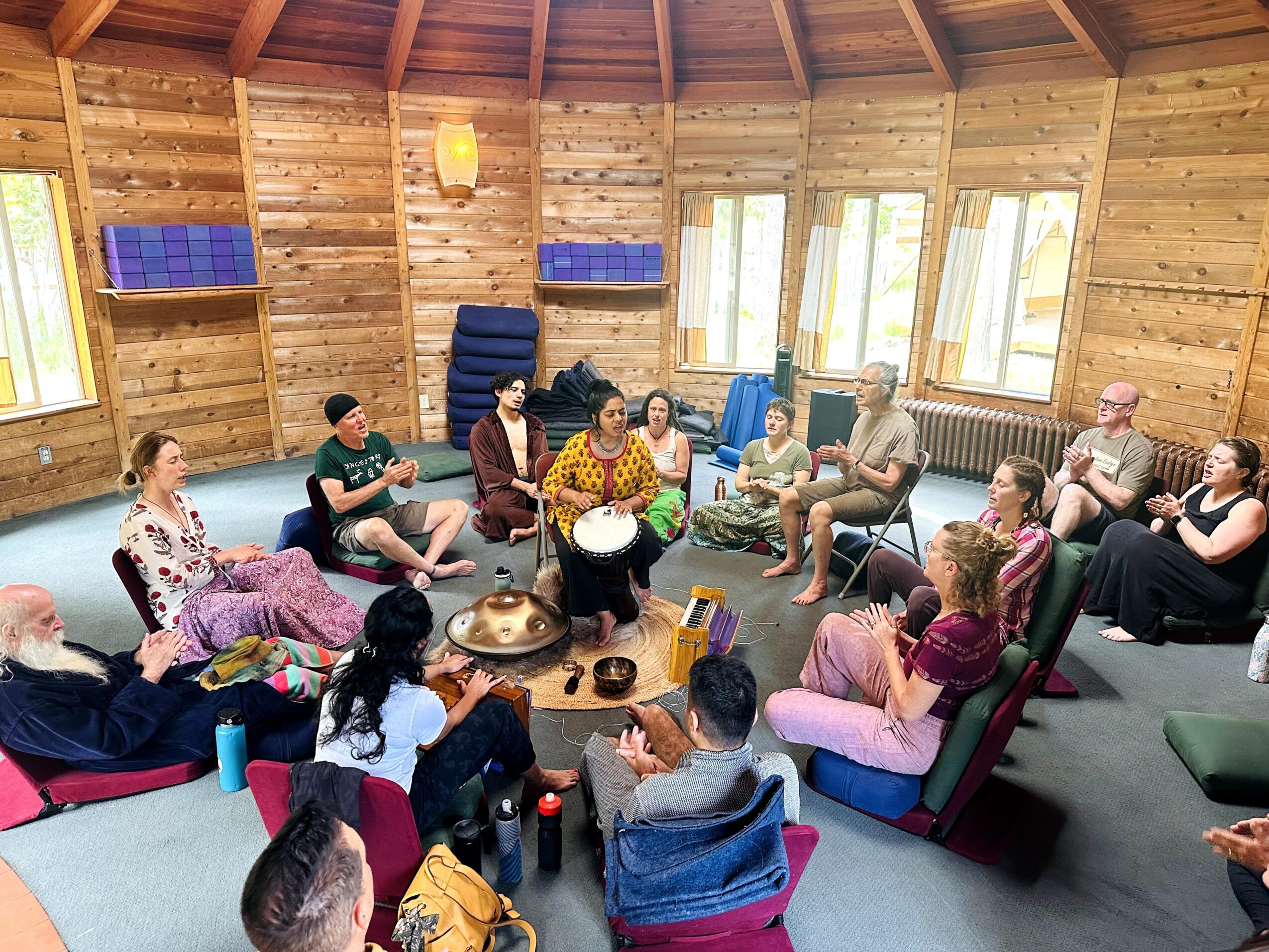 Group of people sitting in a circle inside a wooden room with windows, listening to a person playing a drum.