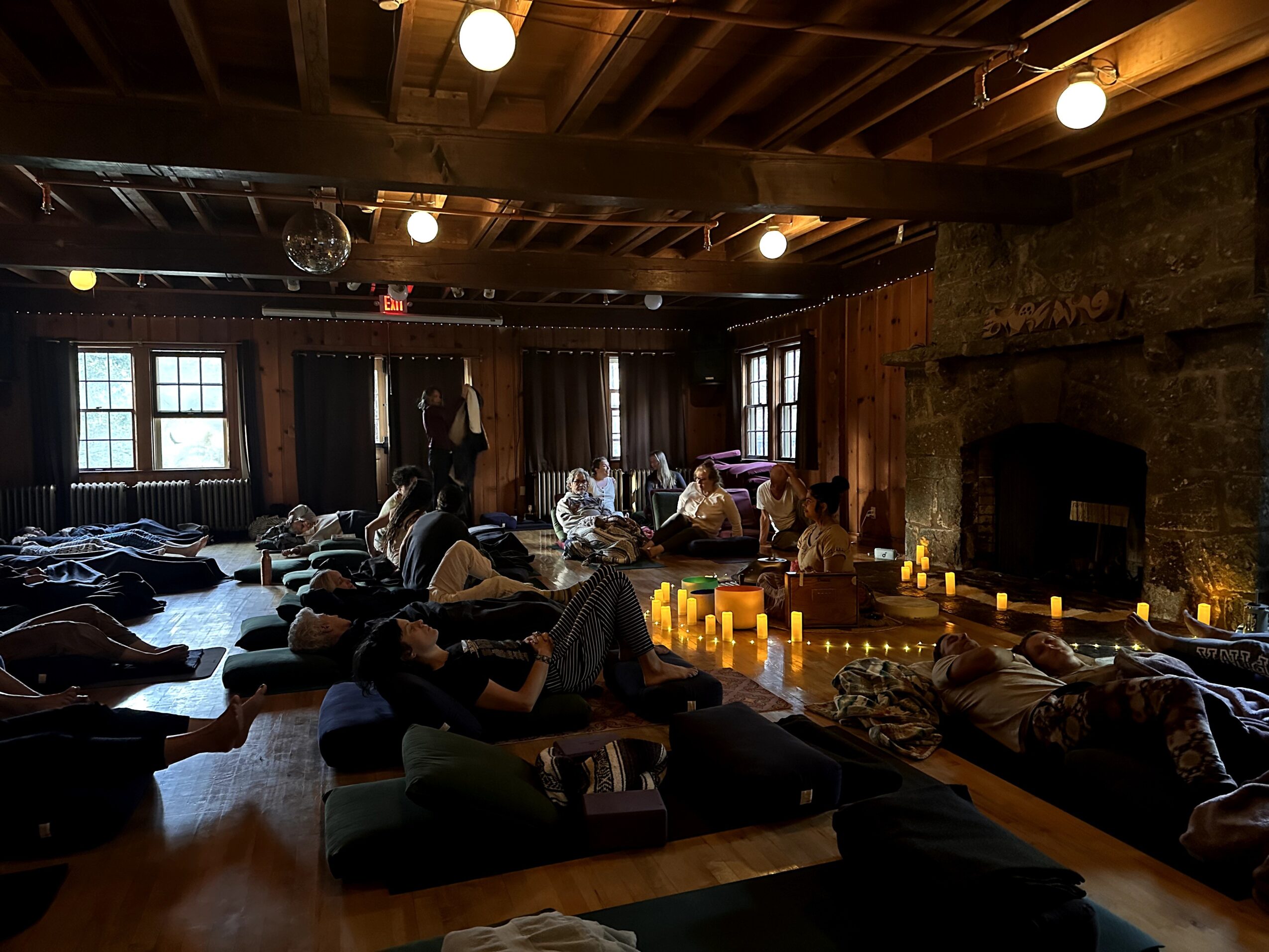People lying on mats in a dimly lit room with candles, wooden walls, and a fireplace, participating in a relaxation session.