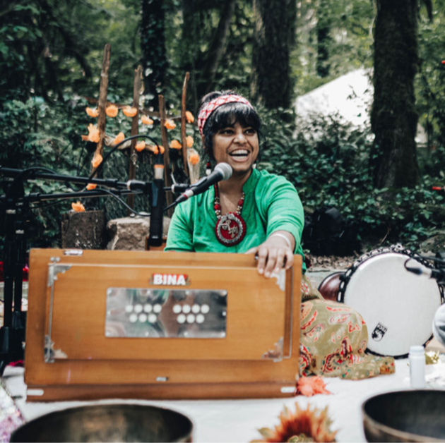 Woman playing a traditional instrument outdoors, smiling, surrounded by trees and musical equipment.