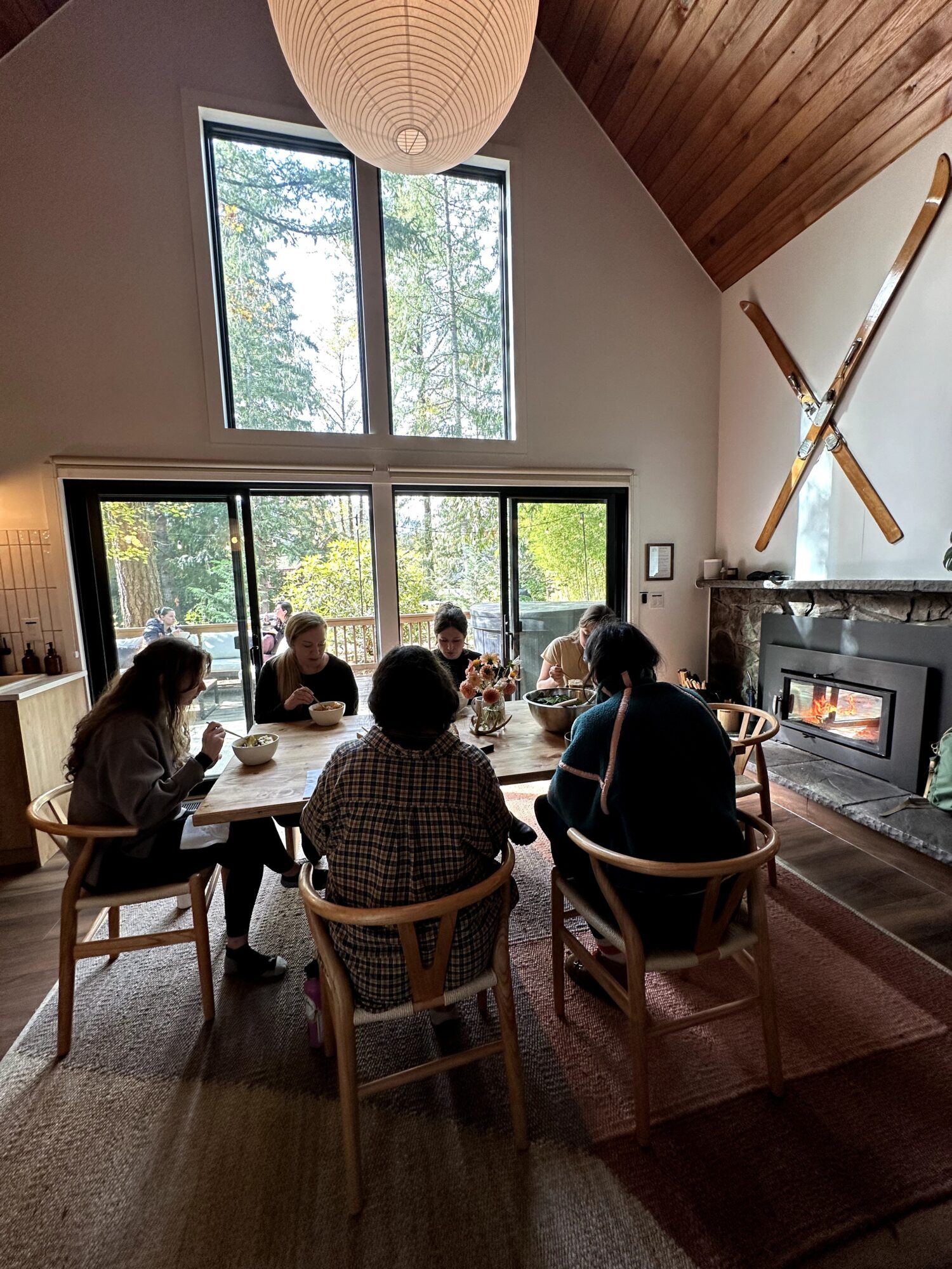 Group of six people sitting around a dining table in a room with large windows, wooden ceiling, and a fireplace.