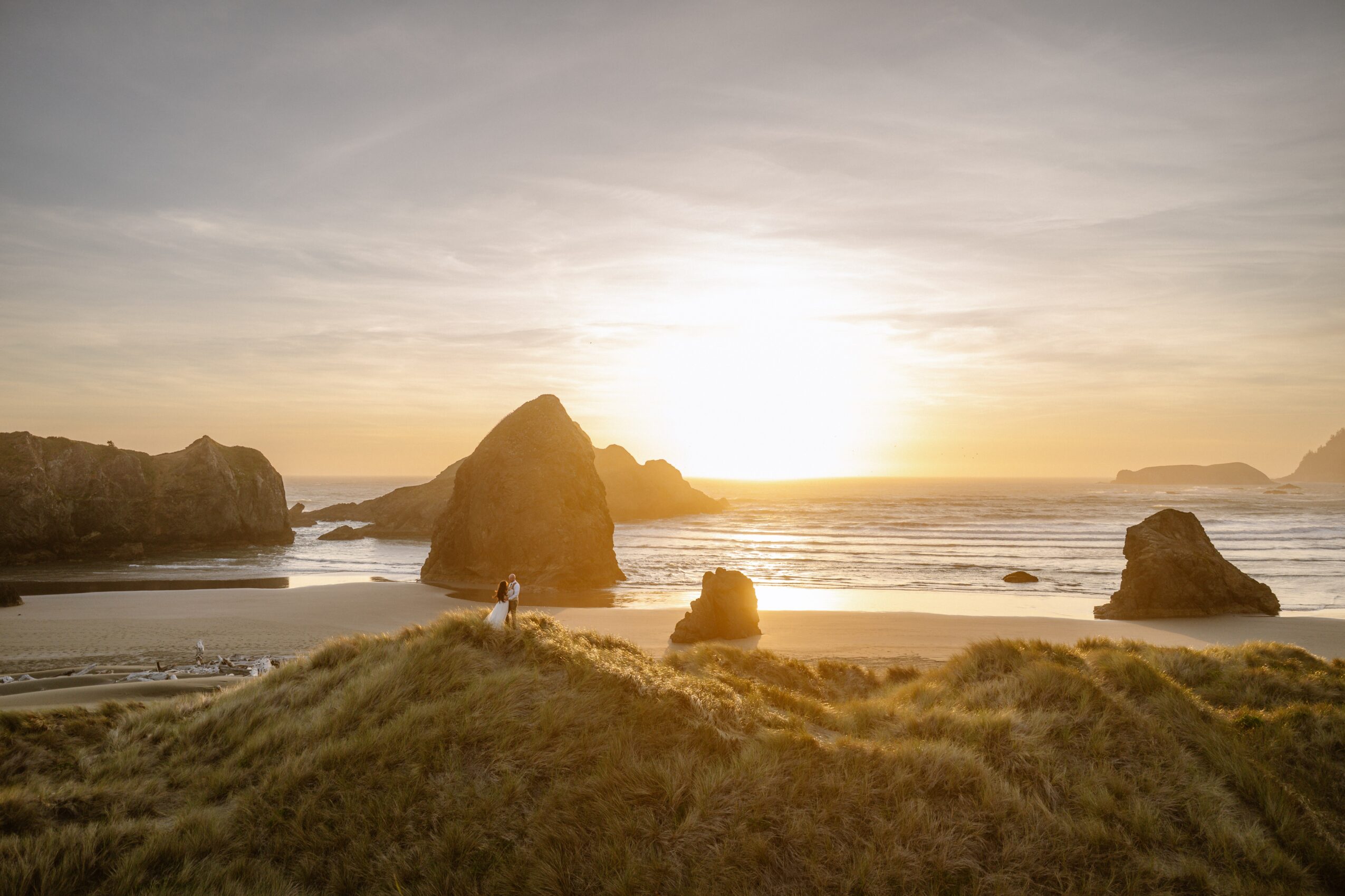 Beach scene with large rock formations, grassy dunes, and a sunset over the ocean, with a person walking along the shore.