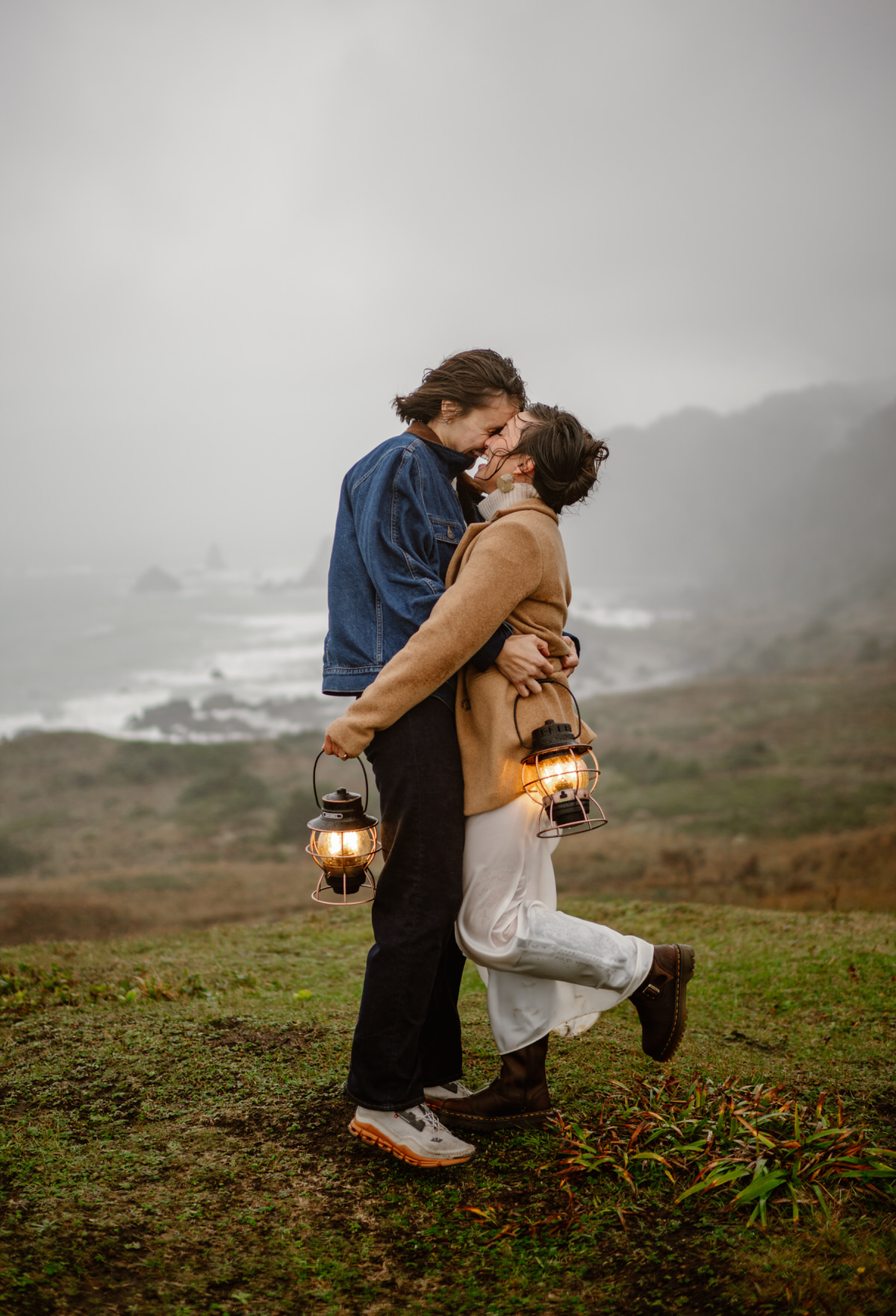A couple embraces outdoors, holding lanterns, with a misty landscape and ocean in the background.