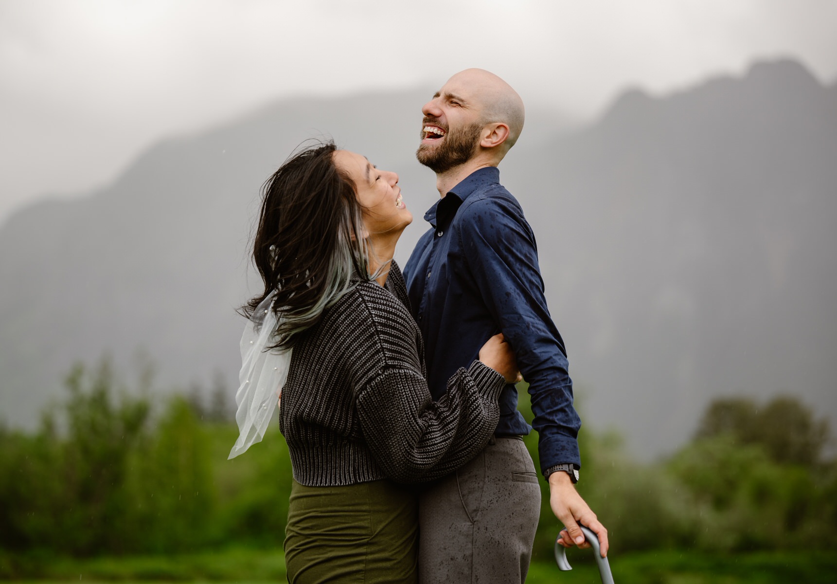 A couple smiling and embracing outdoors with mountains and greenery in the background.