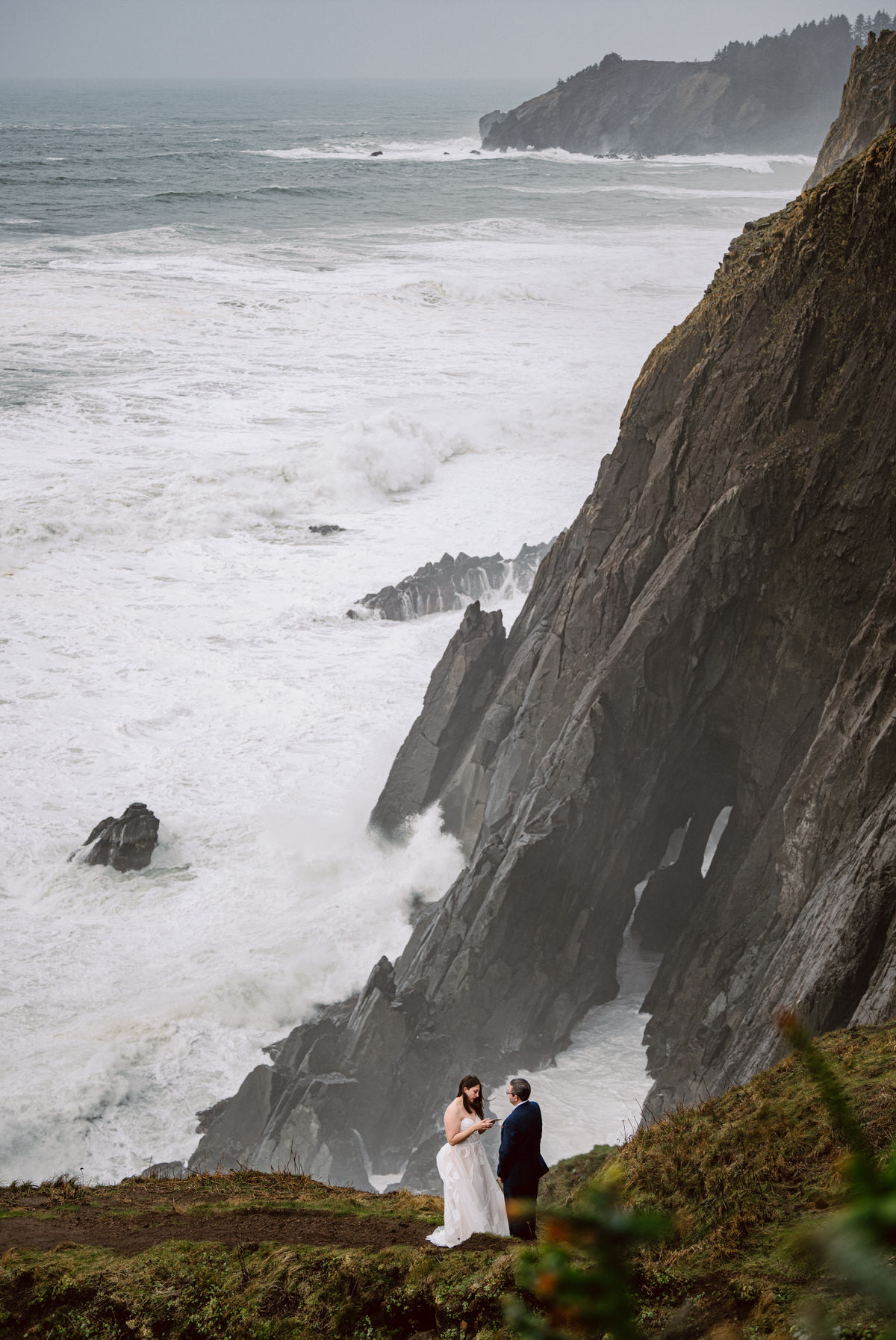A couple stands on a grassy cliff overlooking the ocean with waves crashing against rocks and cliffs in the background.