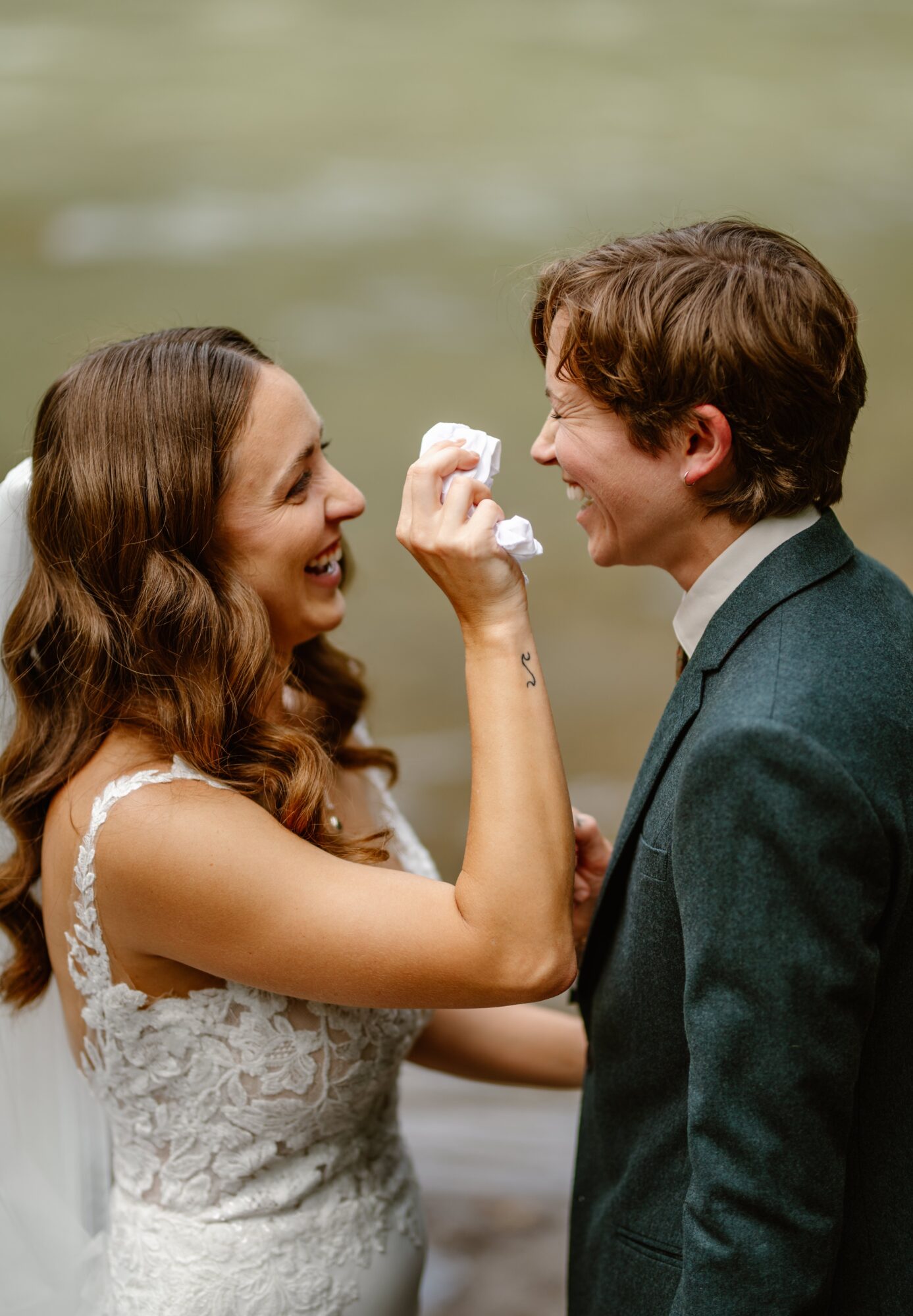 A woman and a man face each other outdoors, smiling, with the woman holding a small object near the man's face.
