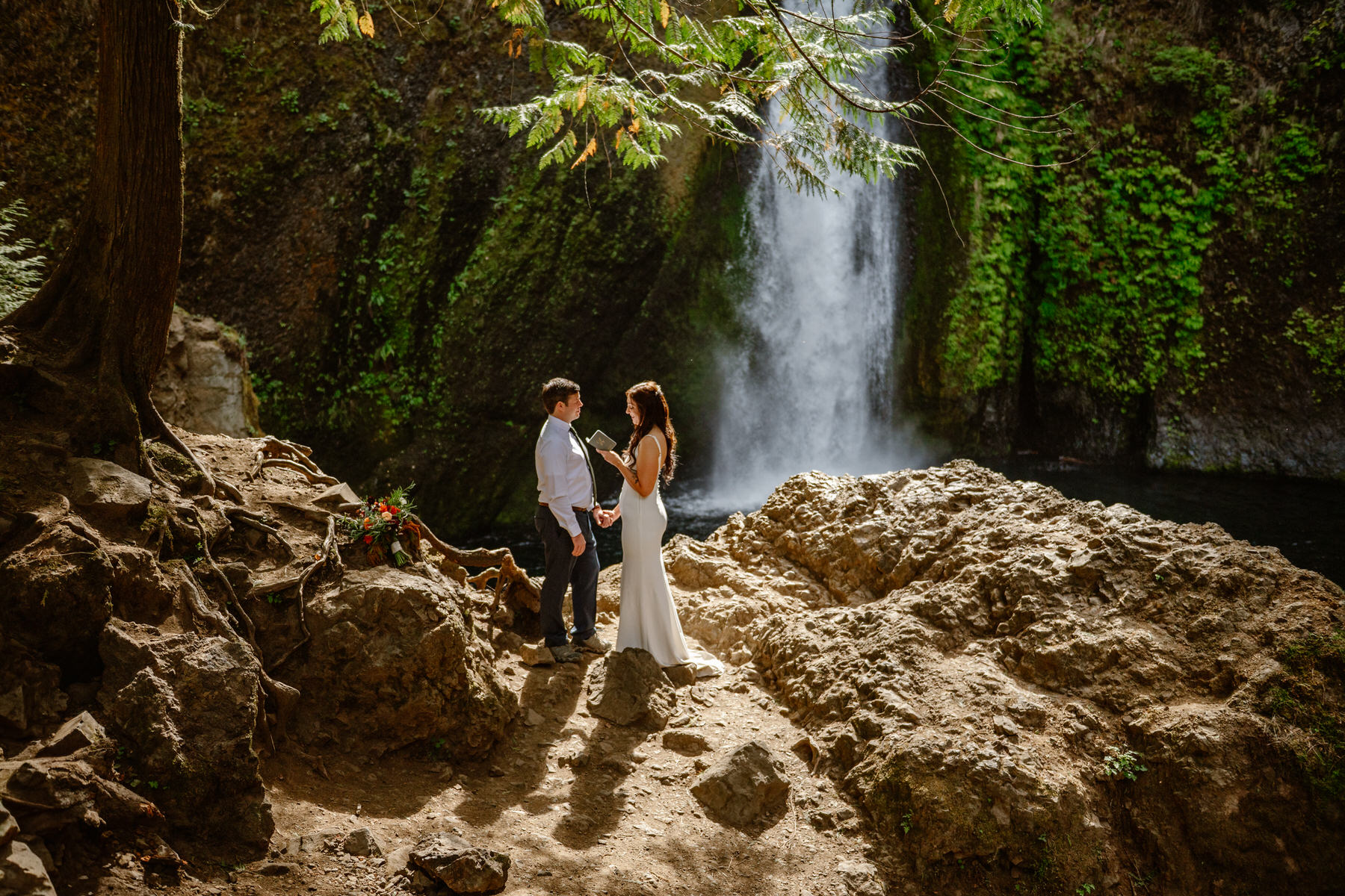 Two people stand on rocks near a waterfall surrounded by greenery, holding hands and facing each other.