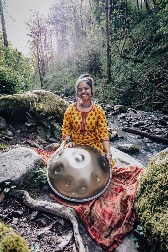 Girl in yellow patterned dress holding a steel pan outdoors near a stream in a forest.