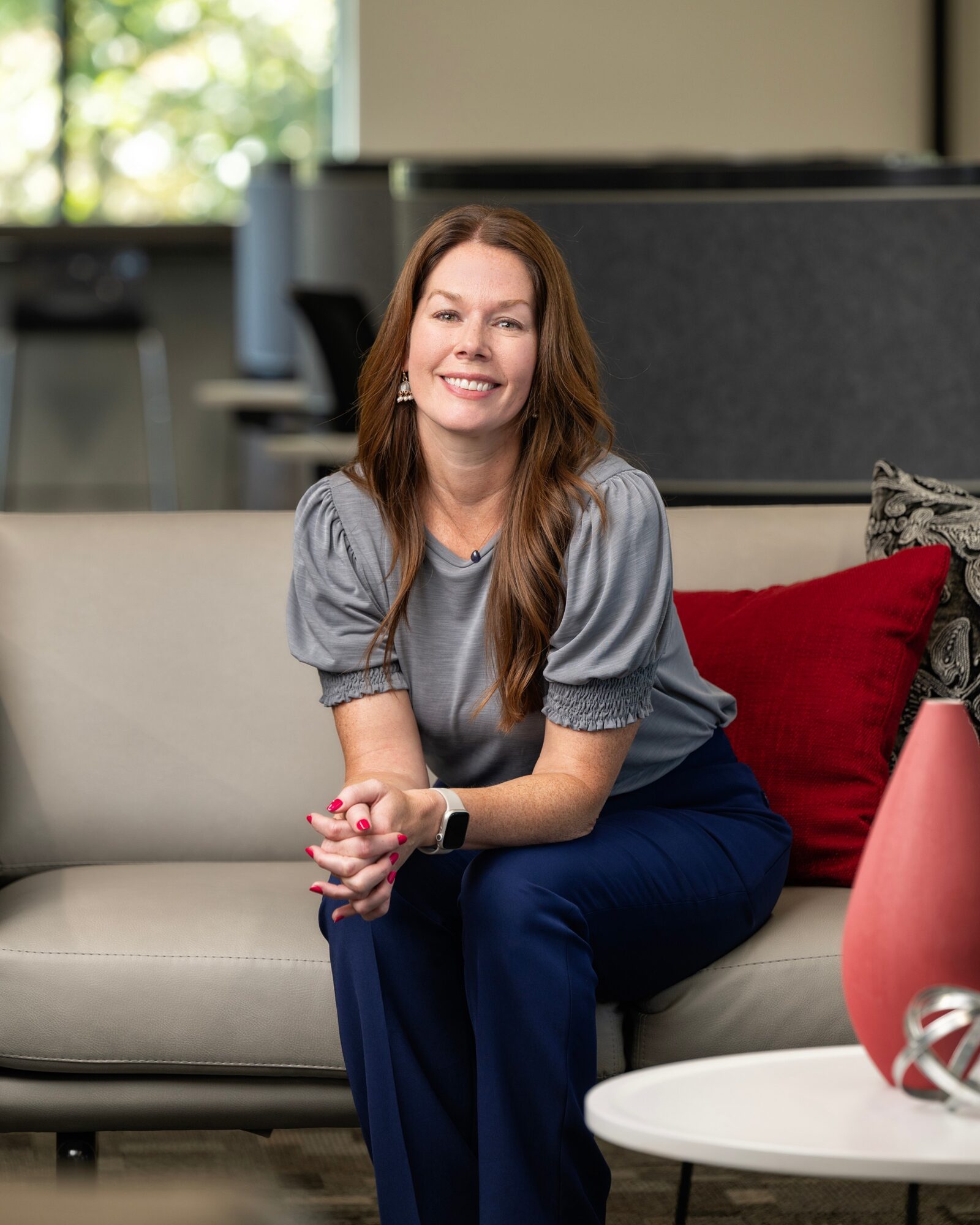Woman with long hair sitting on a sofa, smiling, in a modern living room, with a red pillow and a pink vase nearby.