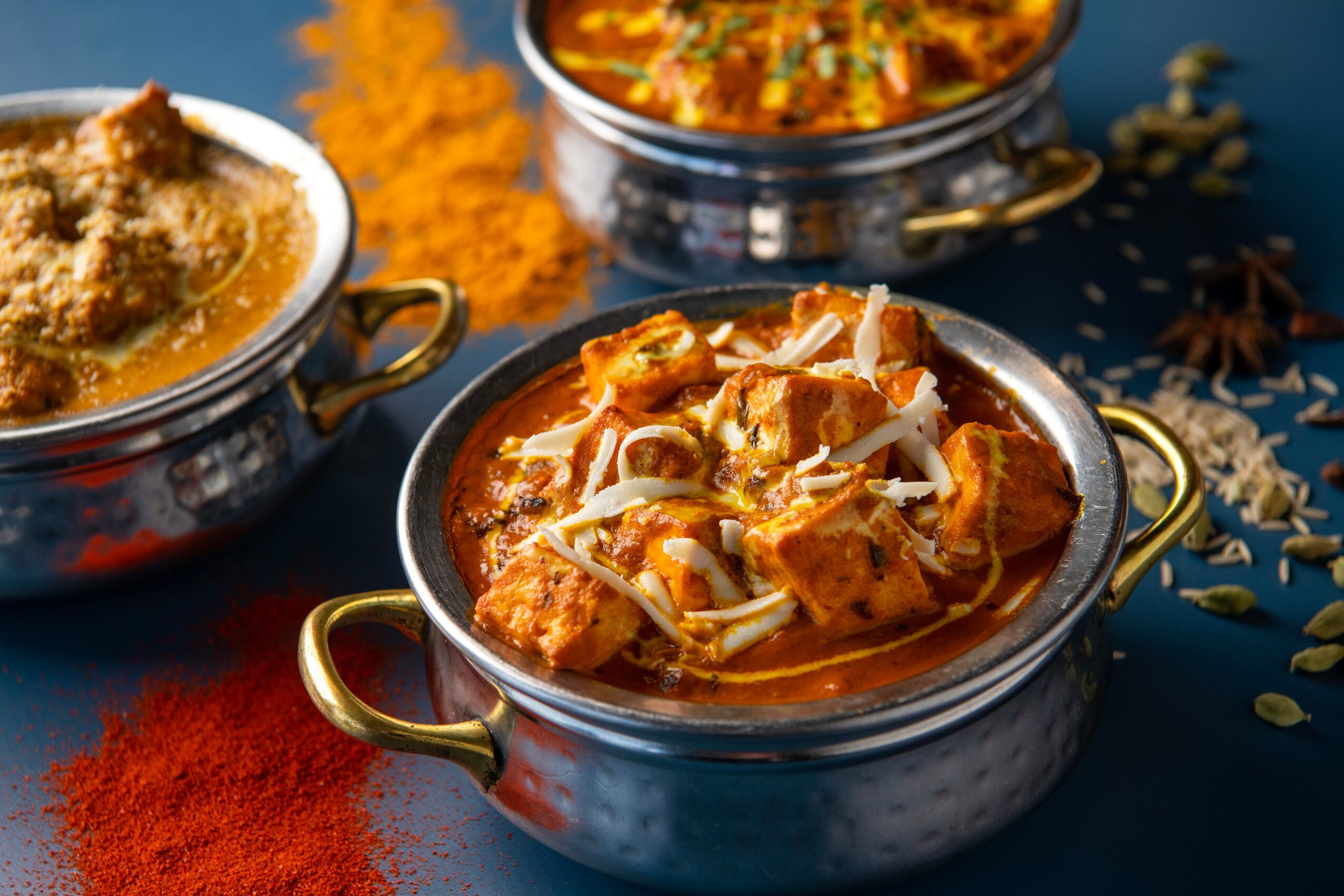 Three bowls of Indian curry dishes with spices and rice, on a dark surface with scattered spices around, colorful background.
