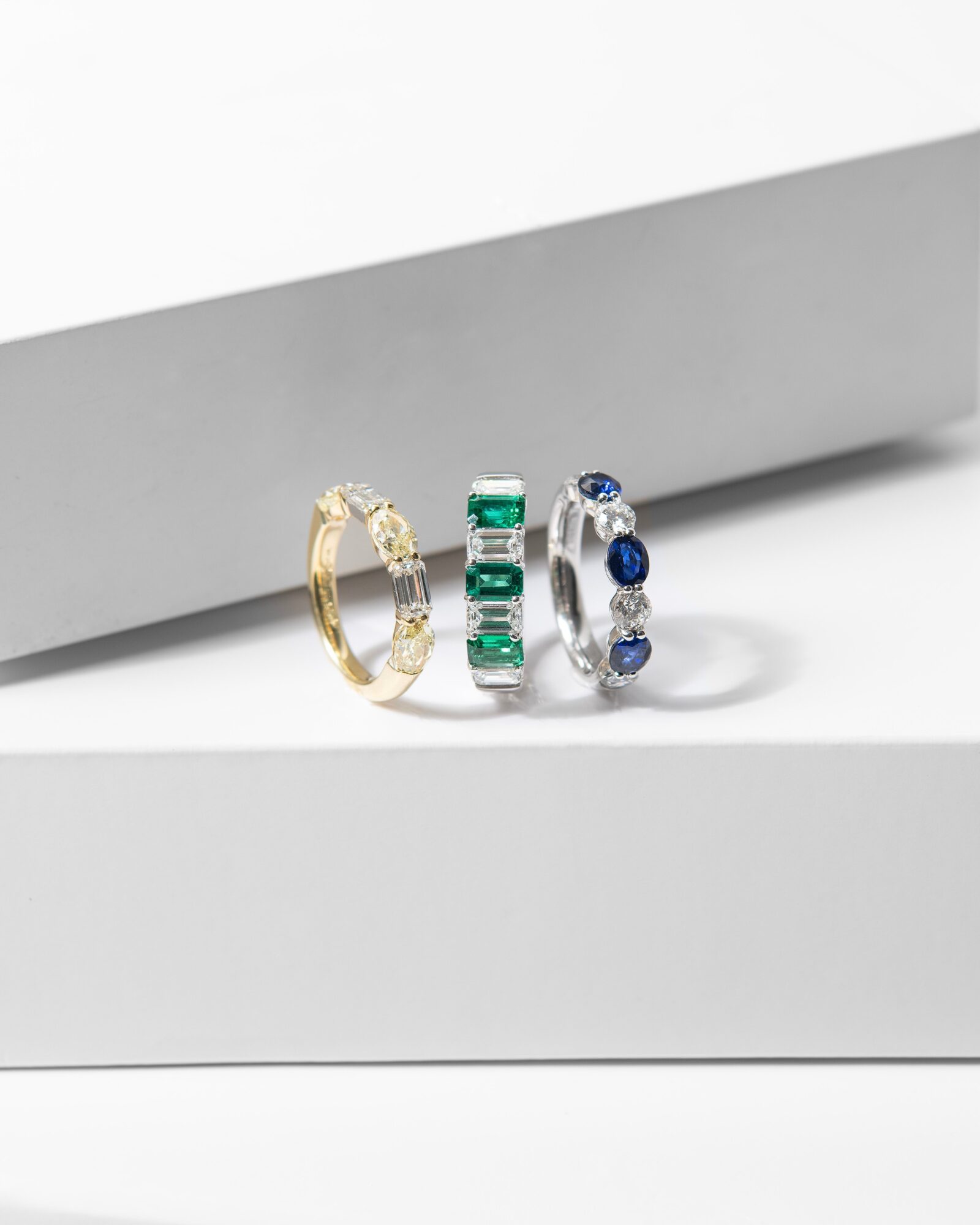 Three rings with gemstones are displayed on a white surface, with a gray background. One gold ring, one with green stones, and one with blue stones.