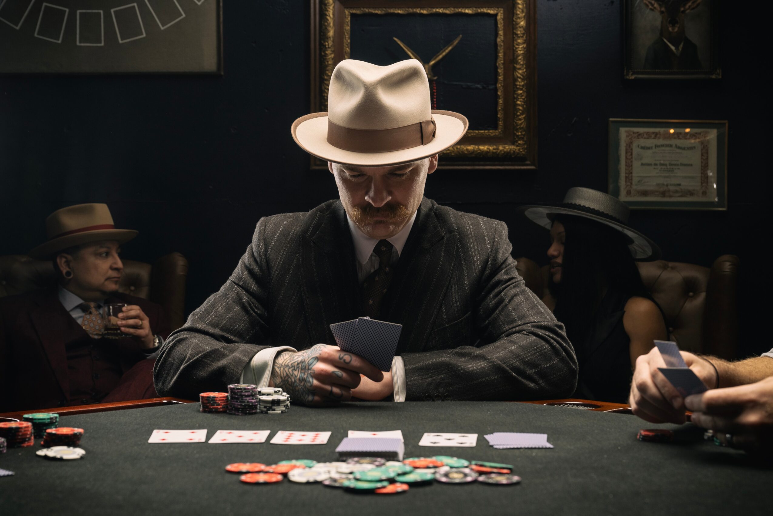 Man in a suit and hat playing poker at a table with chips, with two people in the background, dark room