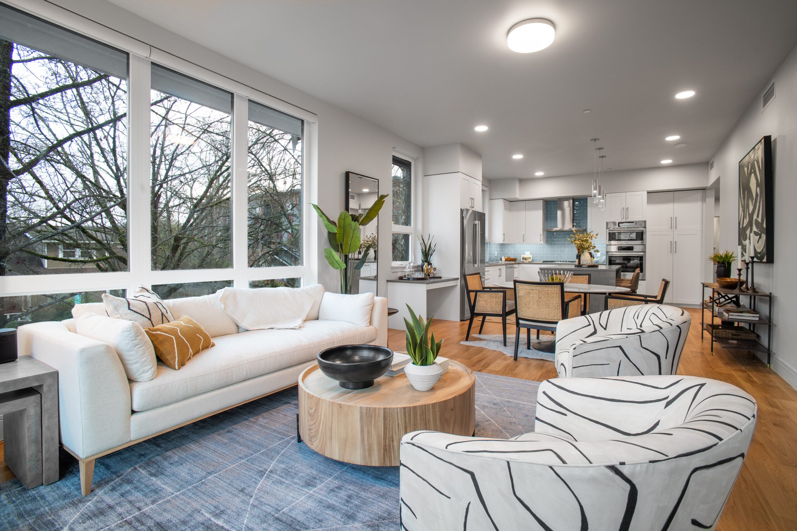 Bright living room with large windows, white sofa, two patterned armchairs, wooden coffee table, and dining area in background.