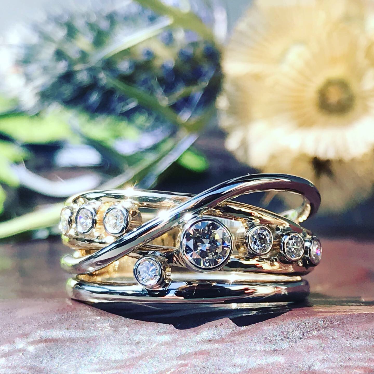Stacked rings with diamonds on a wooden surface, with blurred flowers and greenery in the background.
