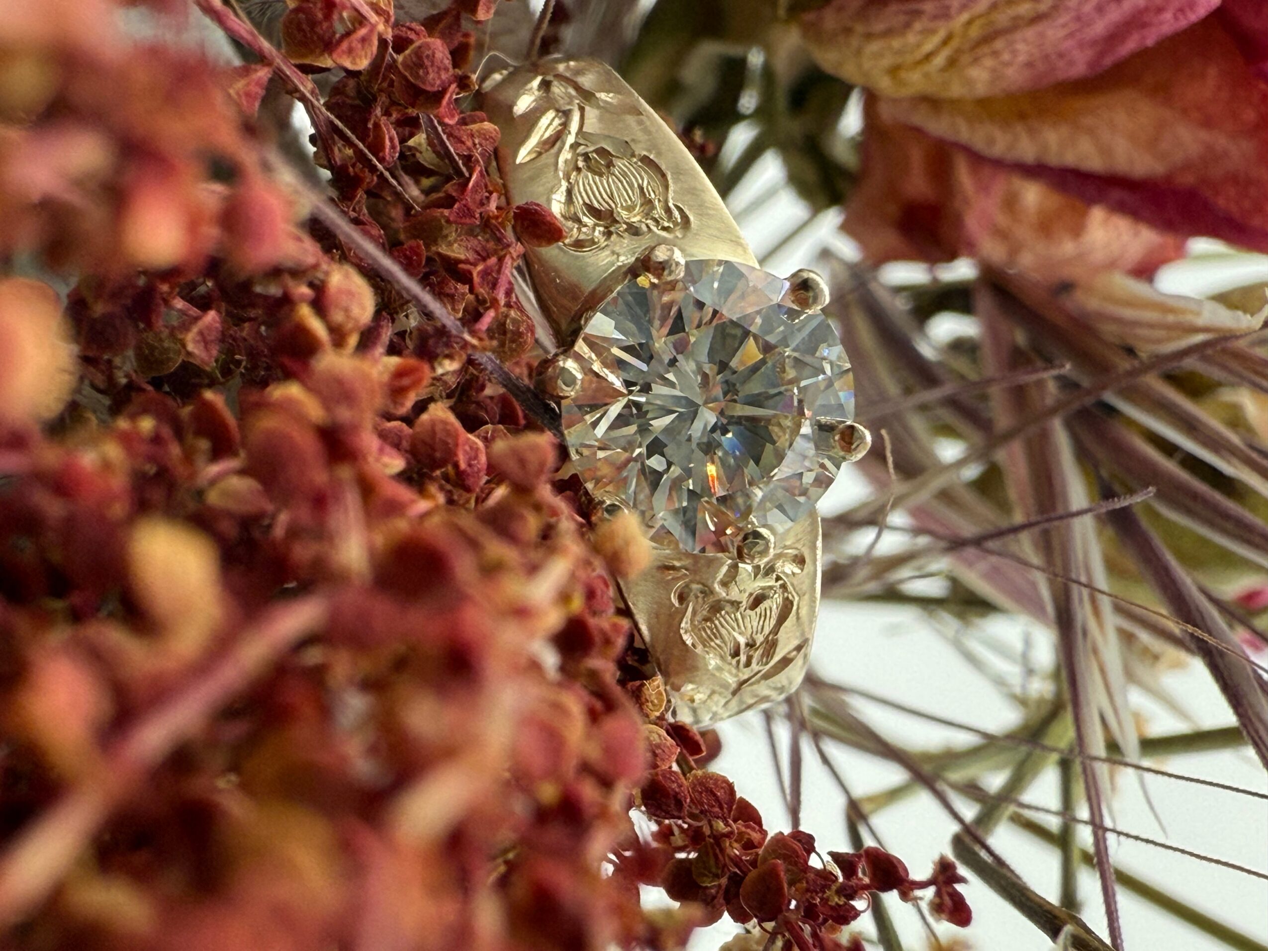 A diamond ring with a large central gemstone surrounded by smaller stones, placed among dried flowers and foliage.