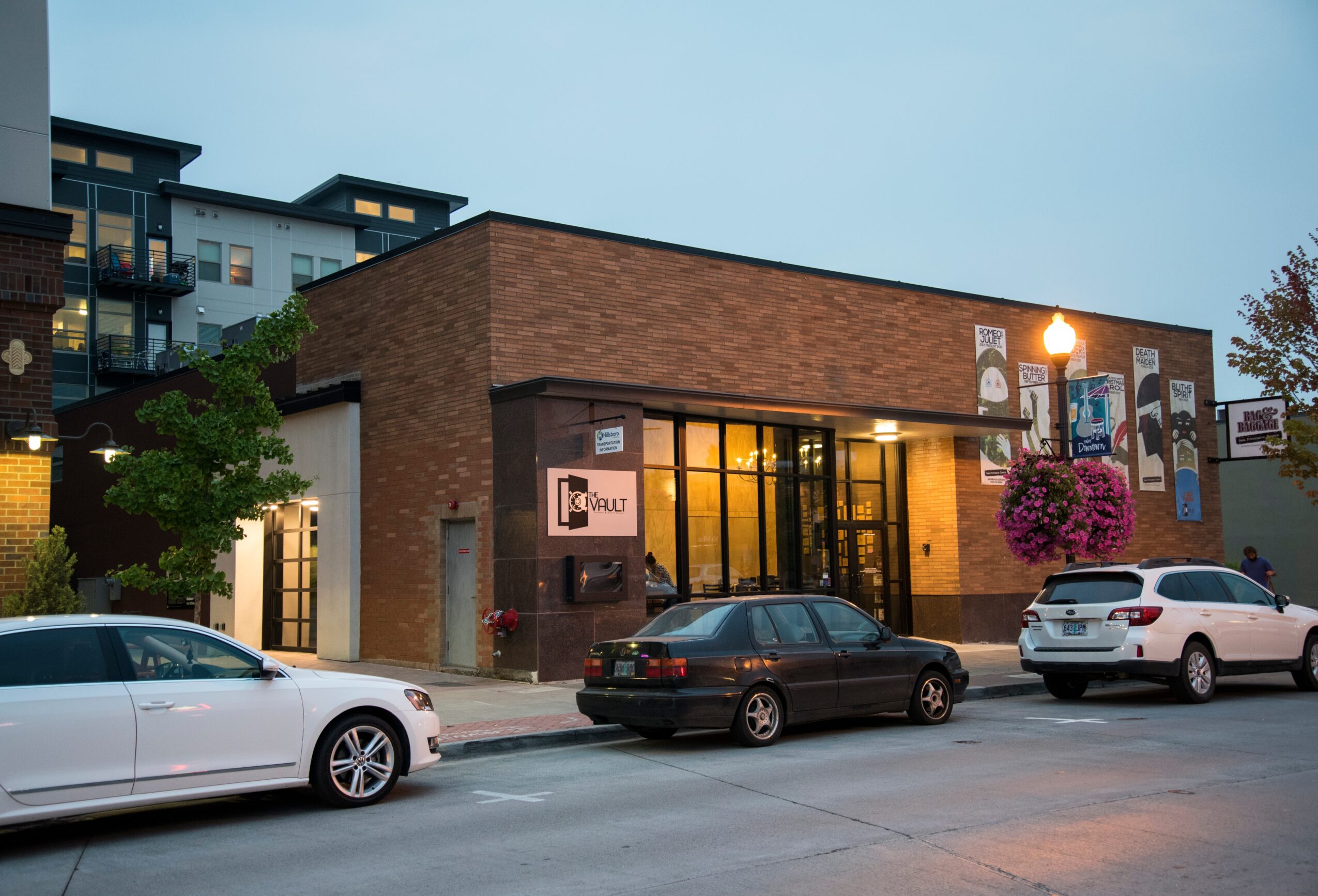 Street view of a brick building with large windows, parked cars, and hanging flower baskets, during evening or dusk.