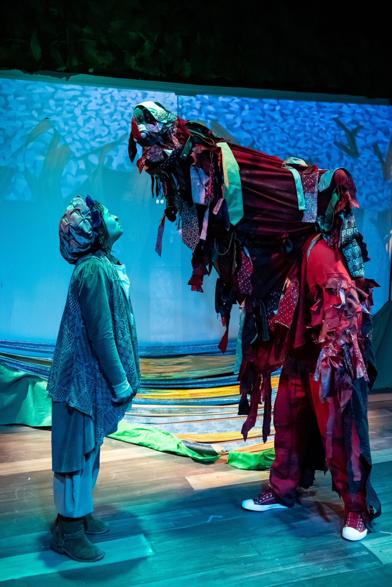 Three performers in colorful costumes and masks on stage, with a large puppet or prop overhead, under blue lighting.
