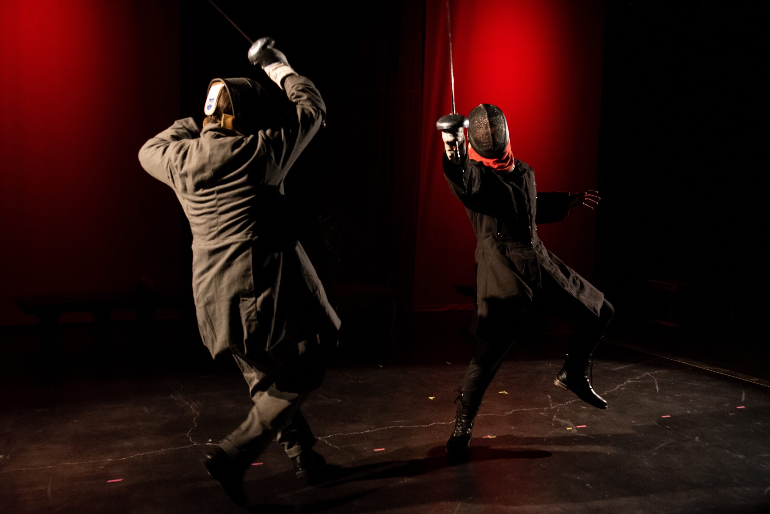 Two performers in black costumes and masks dance on stage with red curtains in the background.