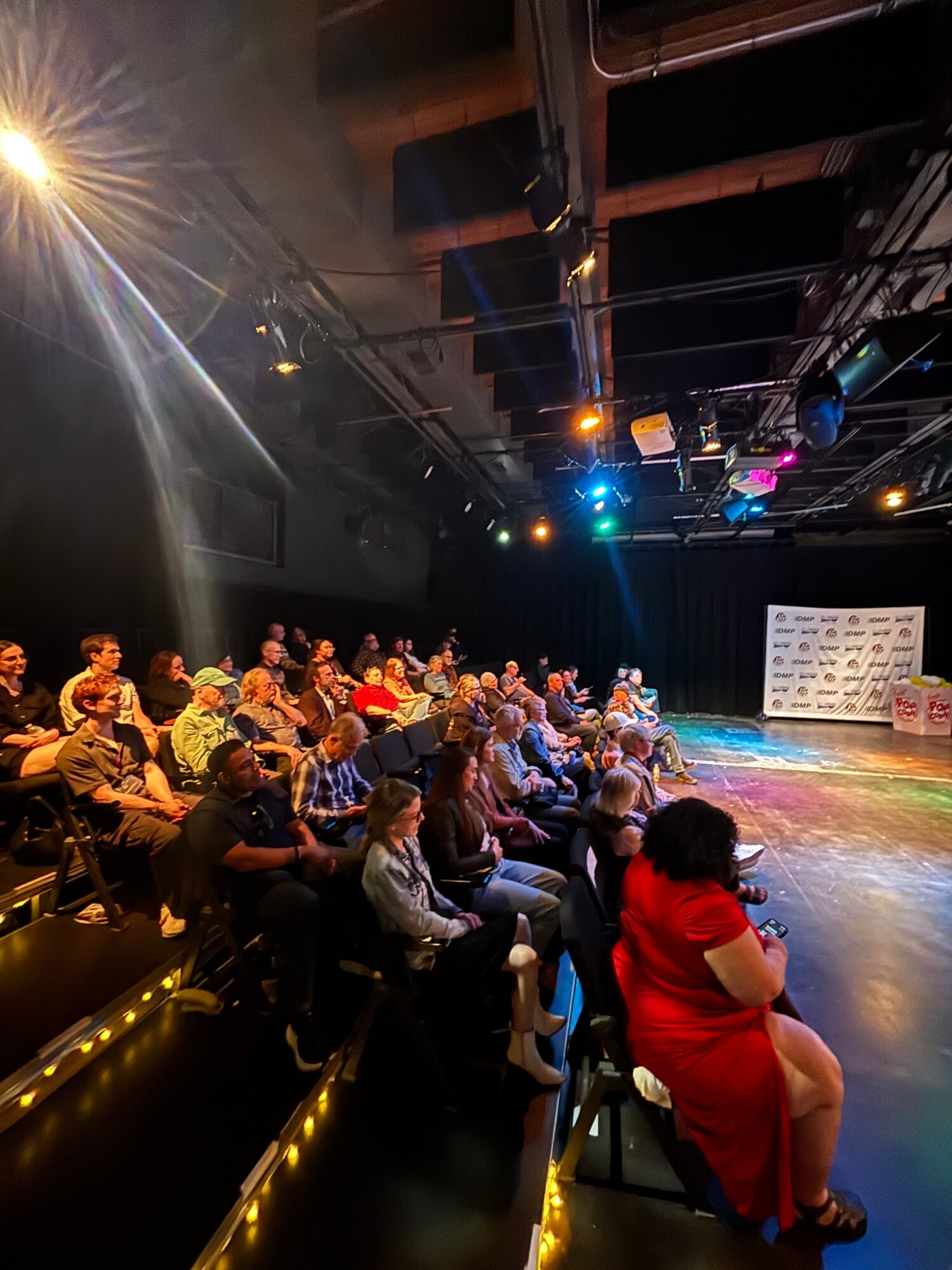 Audience seated in a theater watching a performance on stage with colorful lighting, some people wearing red, black, and white clothing.