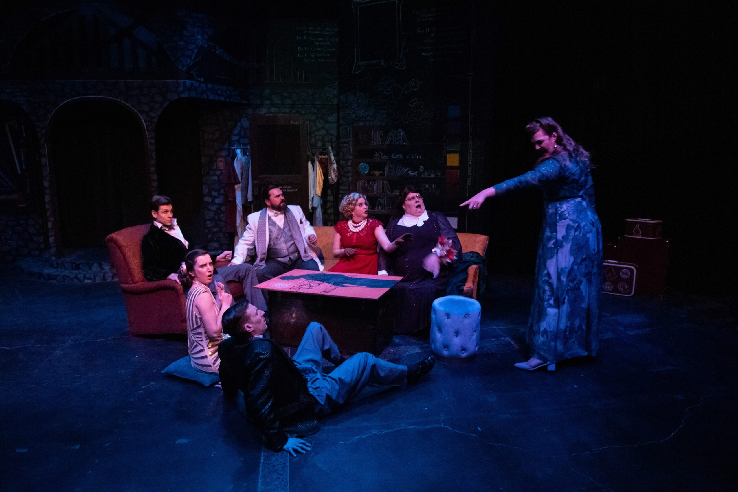 Group of seven people sitting and standing around a table on stage, with a woman standing and gesturing, in a theatrical setting.