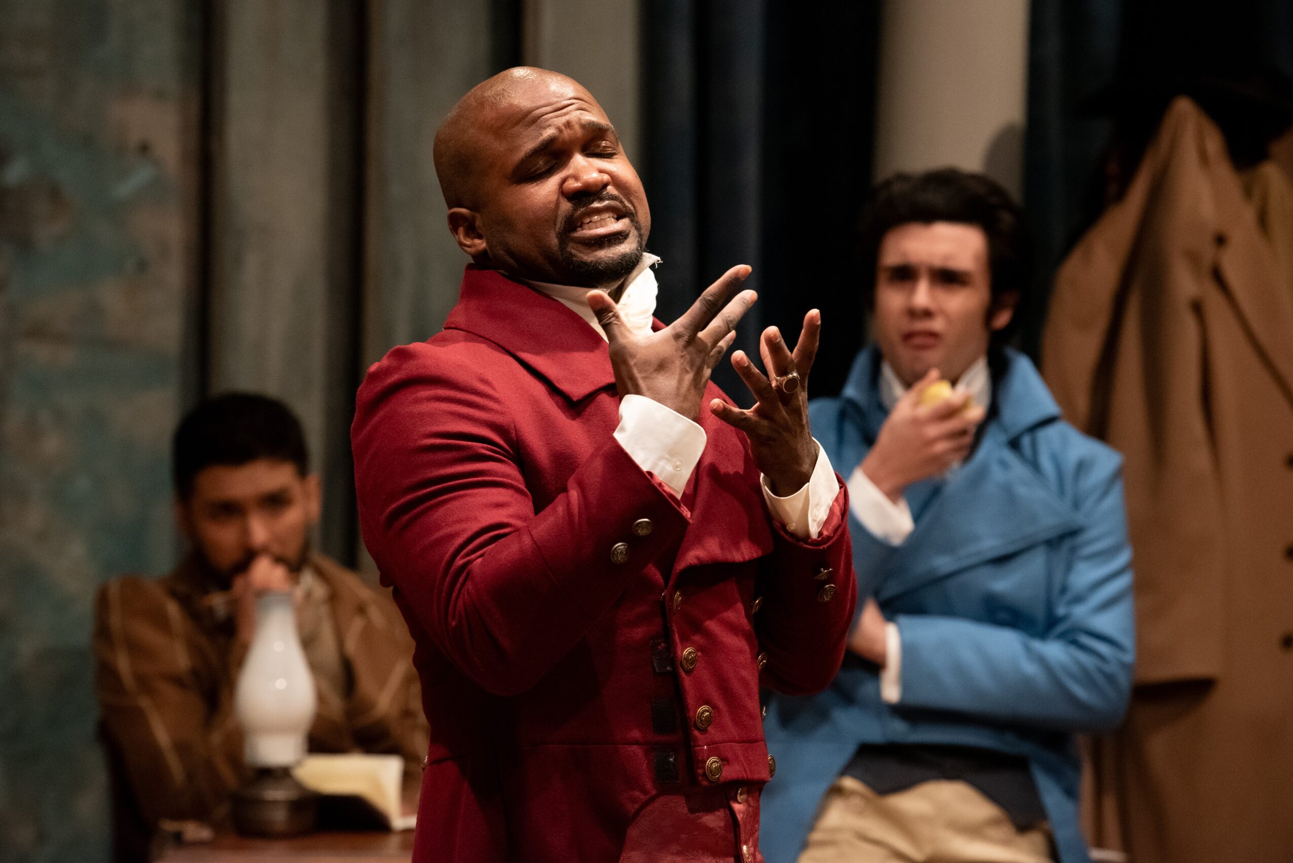 Three men in a room, one in a red jacket speaking passionately, others listening, background with curtains and coat rack.