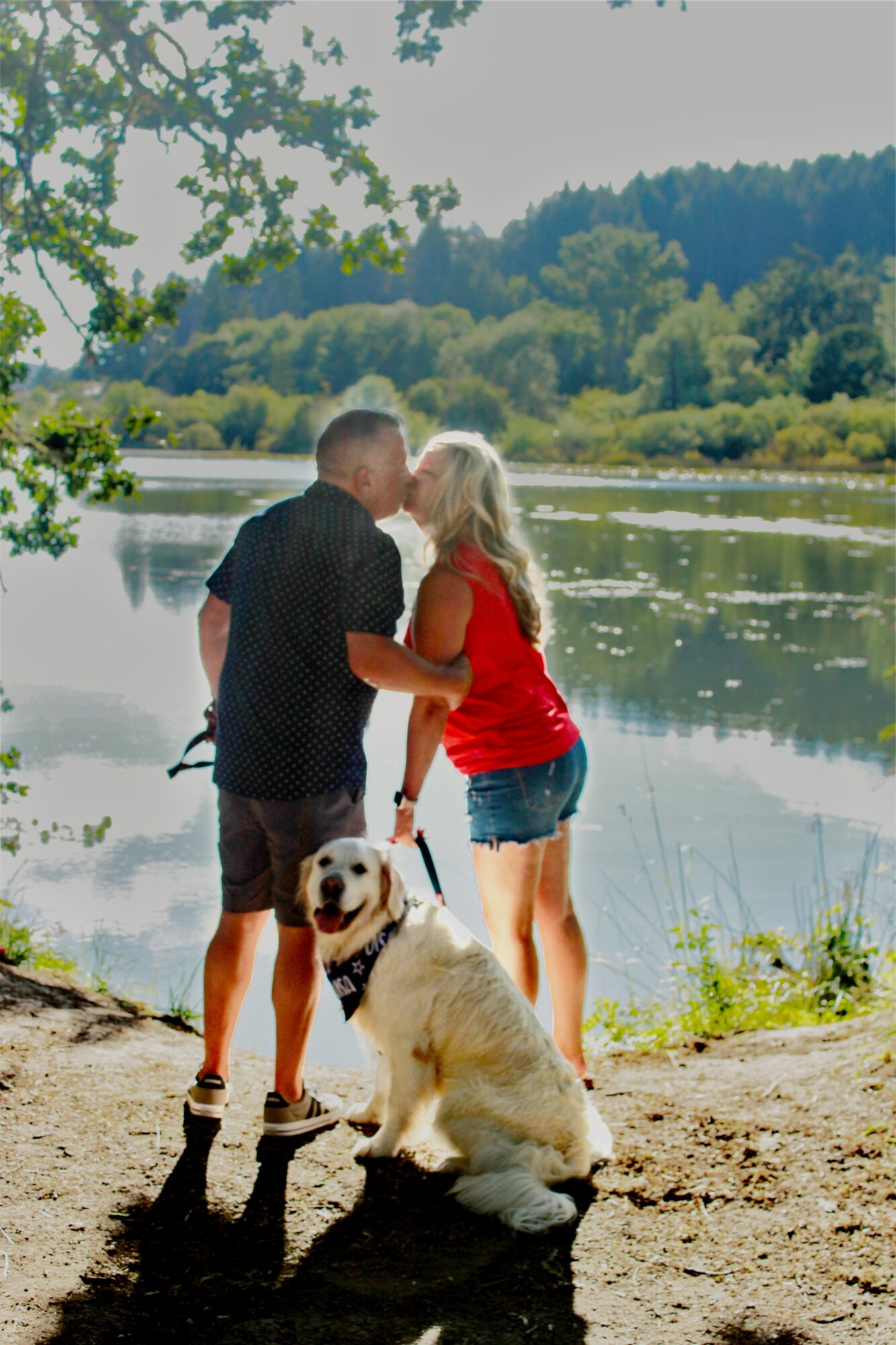 A couple and a dog by a lake with trees and hills in the background, sunny day, couple holding hands, dog sitting.