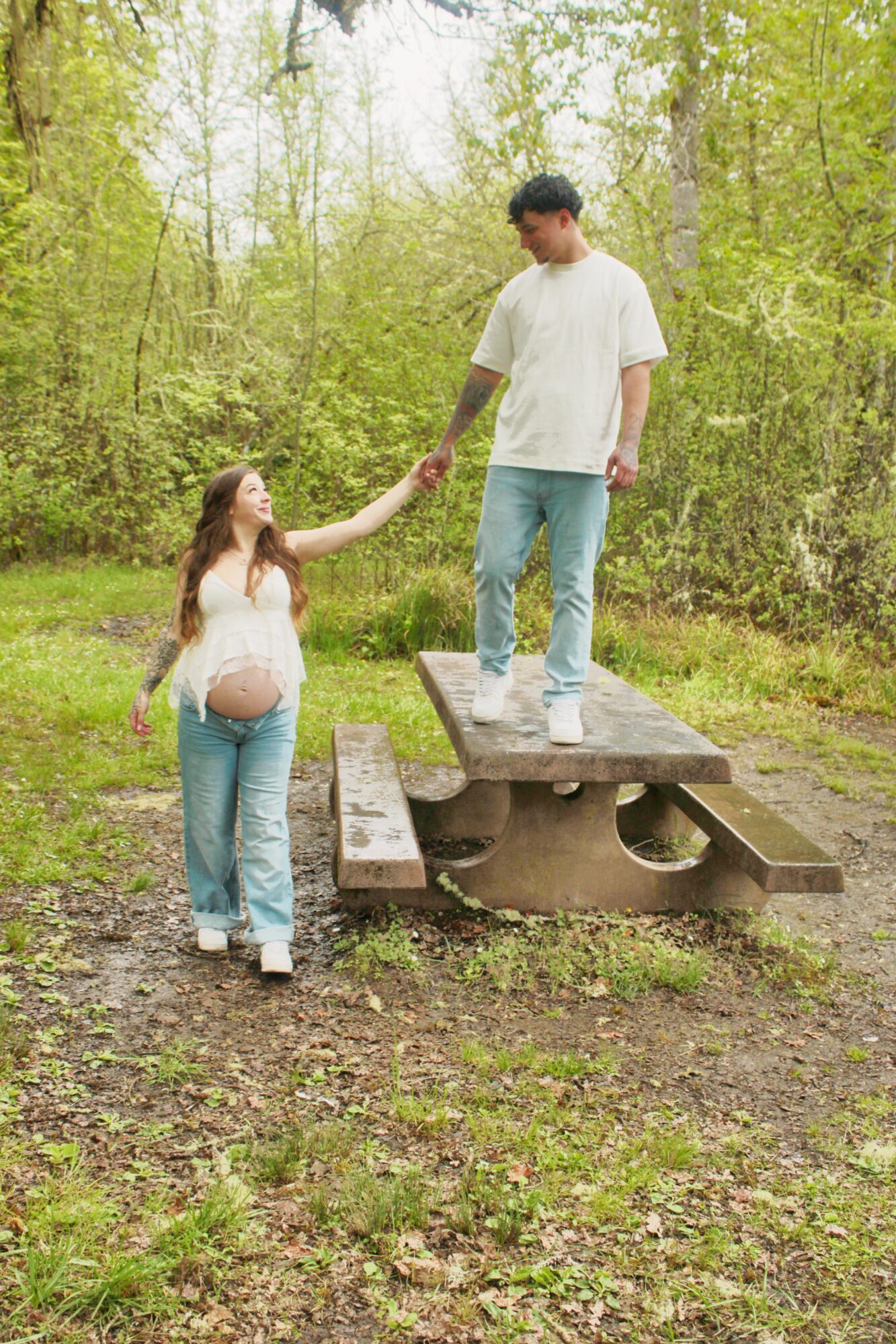 A pregnant woman and a man stand on a picnic table in a forest, holding hands and looking at each other.